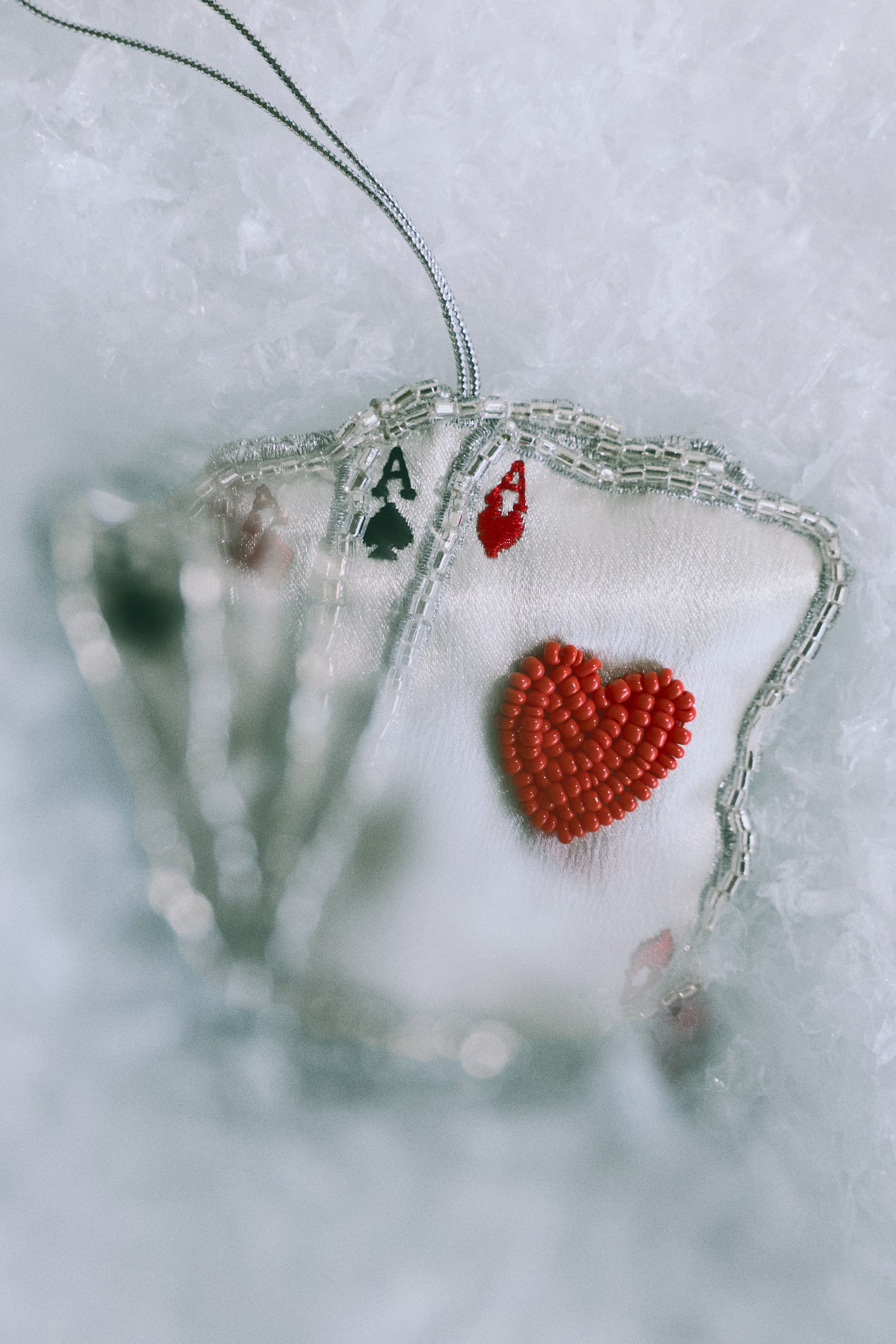 View larger image: A playing card-shaped Christmas ornament is shown from the front, featuring a fan of ace cards crafted from white satin fabric with silver-beaded borders. The front card displays a raised heart made of red beads and an embroidered red 'A' symbol, while a thin silver metallic cord is attached at the top for hanging.