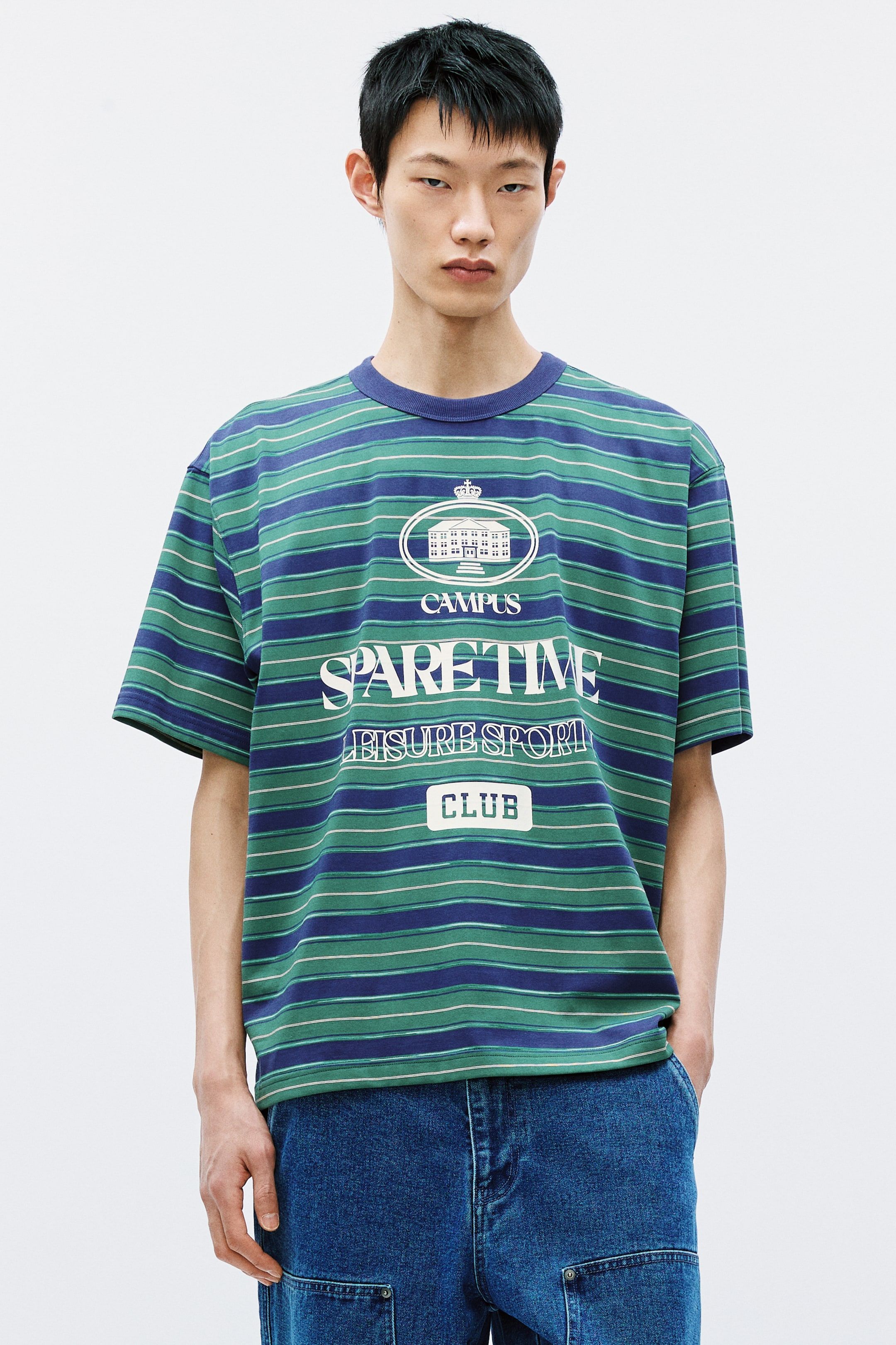 拡大画像を表示: 前面を向いている半袖Tシャツは、ダークブルー、グリーン、細いホワイトの横縞模様が特徴です。フロントには大きなホワイトのグラフィックがあり、建物、クラウン、そして「CAMPUS SPARE TIME LEISURE SPORT CLUB」というテキストが含まれています。また、ダークブルーのクルーネックカラーを備えています。