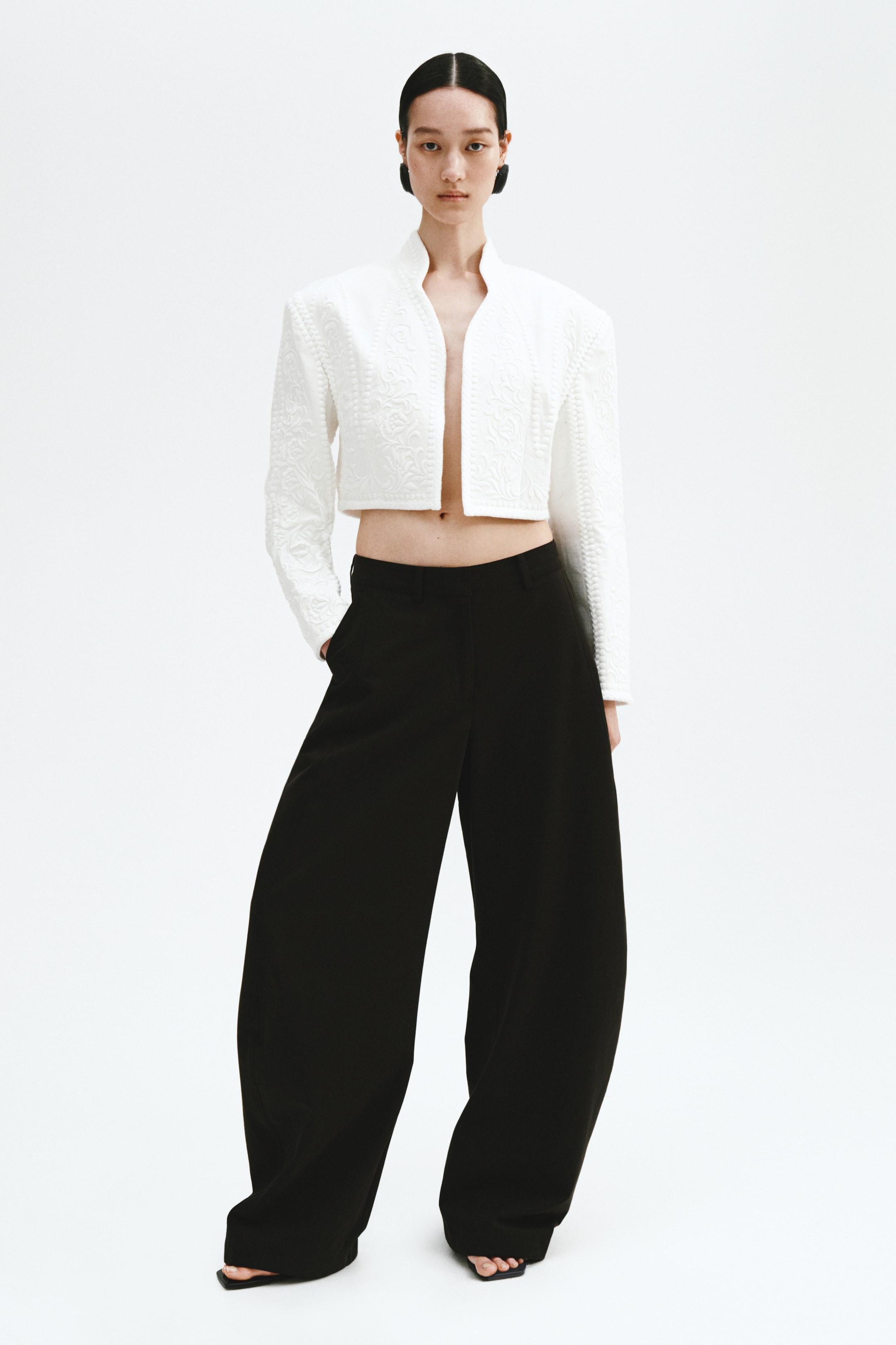 View larger image: A white cropped jacket showcasing a standing collar, structured shoulders, and a textured floral pattern from the front view. It's styled open over black high-waisted, wide-leg trousers, with one hand casually placed in a trouser side pocket. The full-length hems of the trousers elegantly drape over the feet.