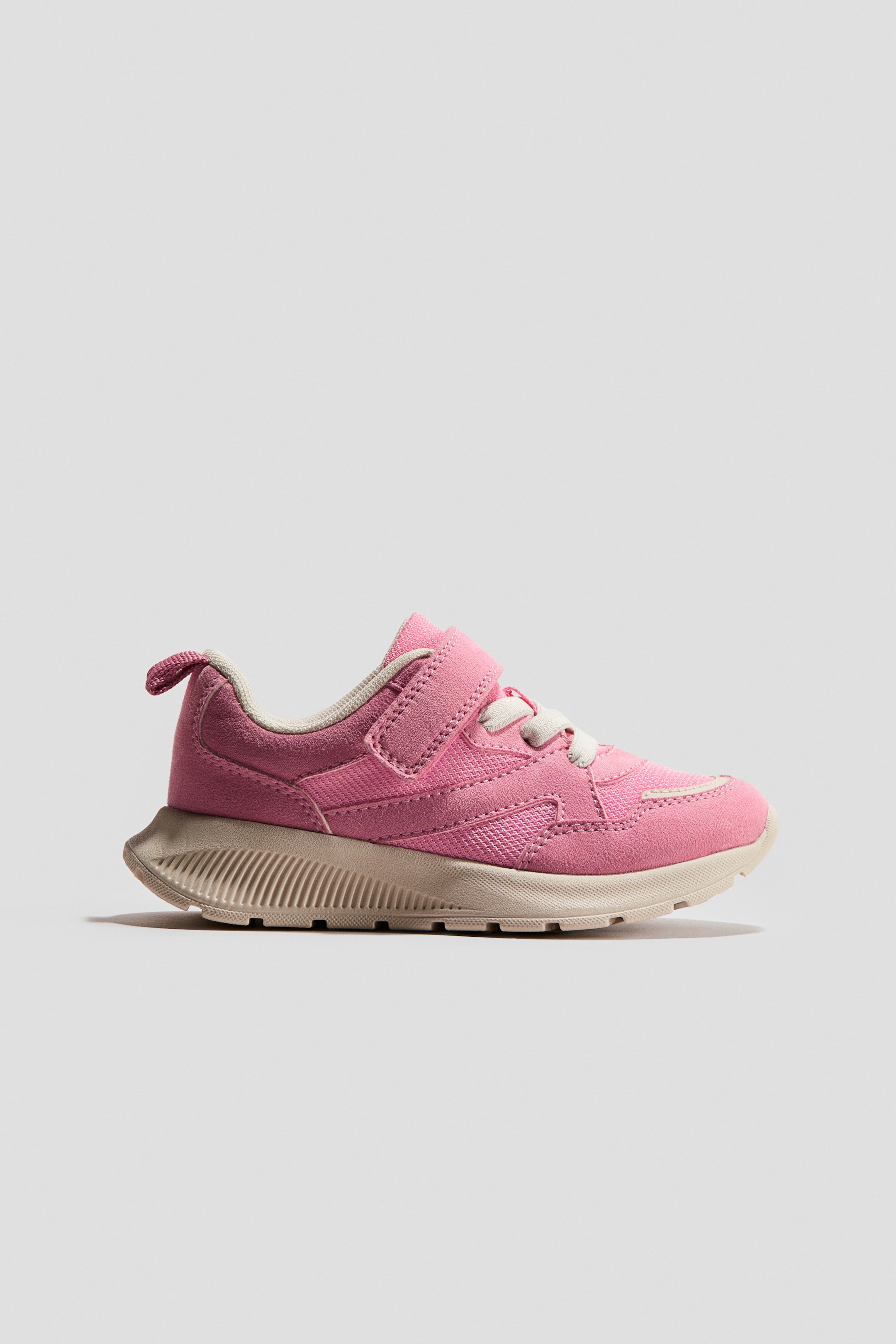 Visualizza immagine più grande: Una sneaker rosa presenta il suo profilo laterale, caratterizzato da una tomaia rosa con pannelli in mesh e simil camoscio, e una piccola linguetta per il tallone si estende dal retro. Una strap in velcro copre i lacci elastici, e la scarpa poggia su una suola chunky di colore beige chiaro che include una trama a coste nel midsole.