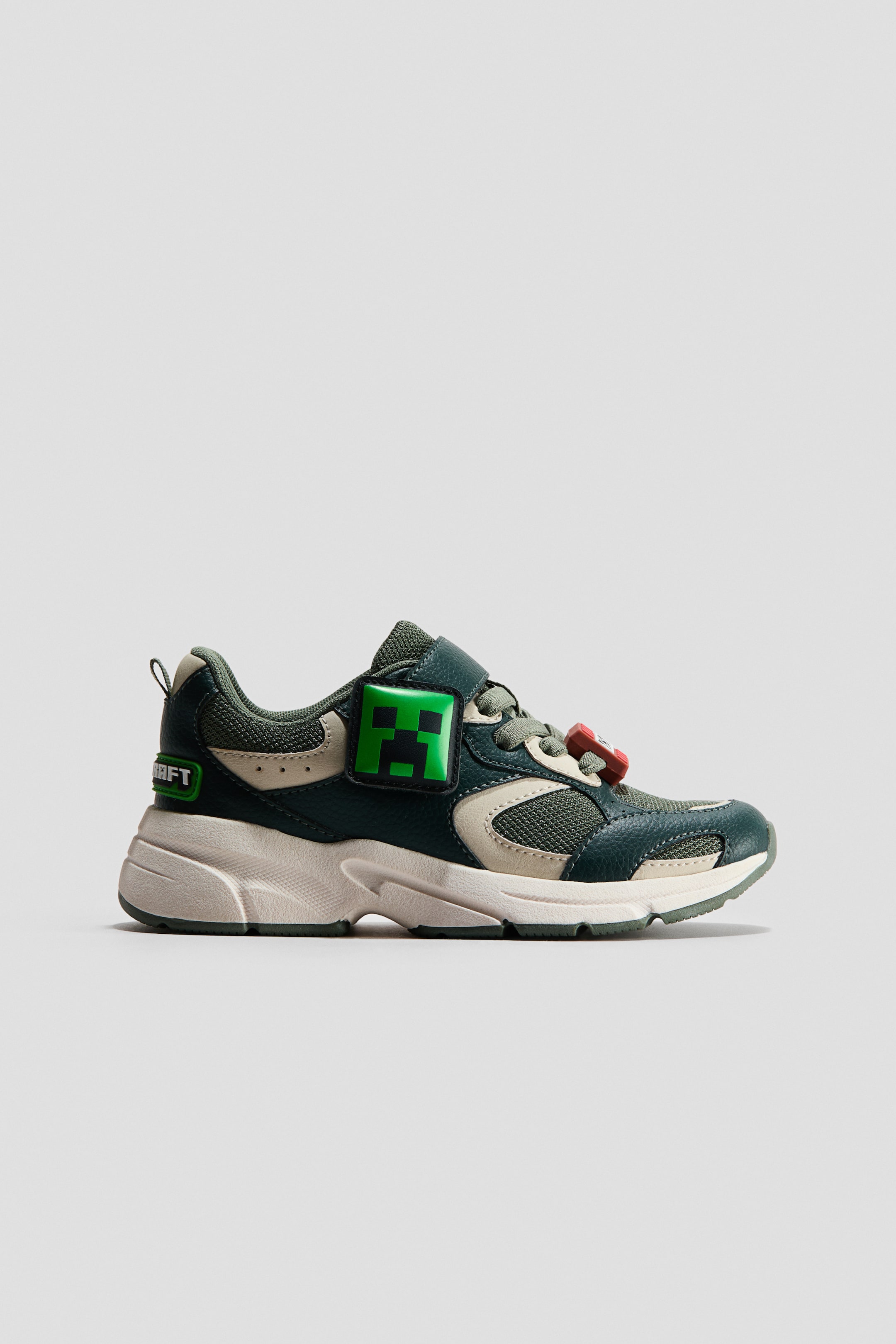 View larger image: A dark green and beige trainer is facing left, displaying mesh and synthetic leather panels. It features a velcro strap with a green pixelated Creeper face, a small red pixelated charm on the laces, the word 'CRAFT' on the heel, and a chunky cream sole.
