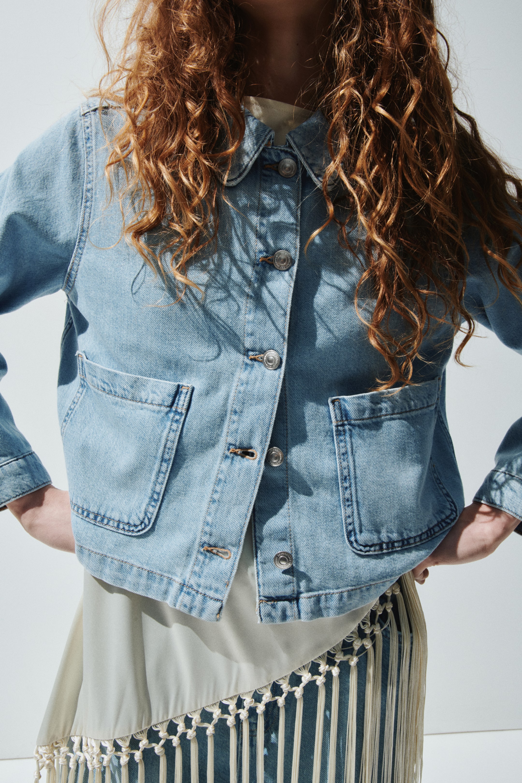 View larger image: The light blue denim jacket features a pointed collar and a silver-toned button-down front. It also includes two large rounded patch pockets with contrasting orange topstitching, and is styled partially unbuttoned over a light-coloured garment.