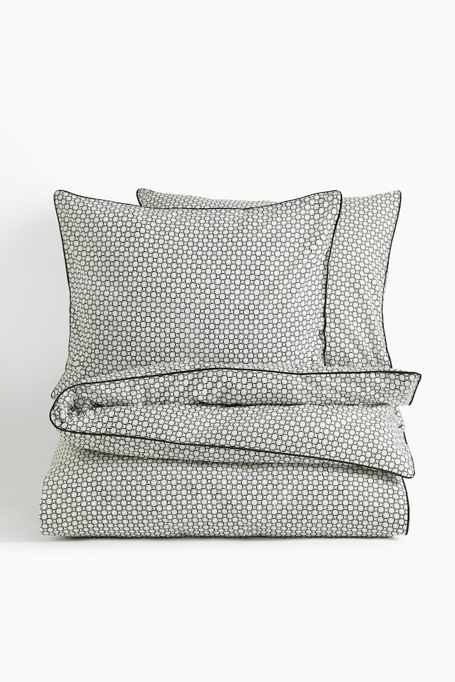A bedding set is featuring a neatly folded duvet cover and two upright pillowcases, all showcasing a small, repeating black-and-white geometric grid pattern. Each item is displaying dark green piped edges, with the set arranged facing the front. - 1