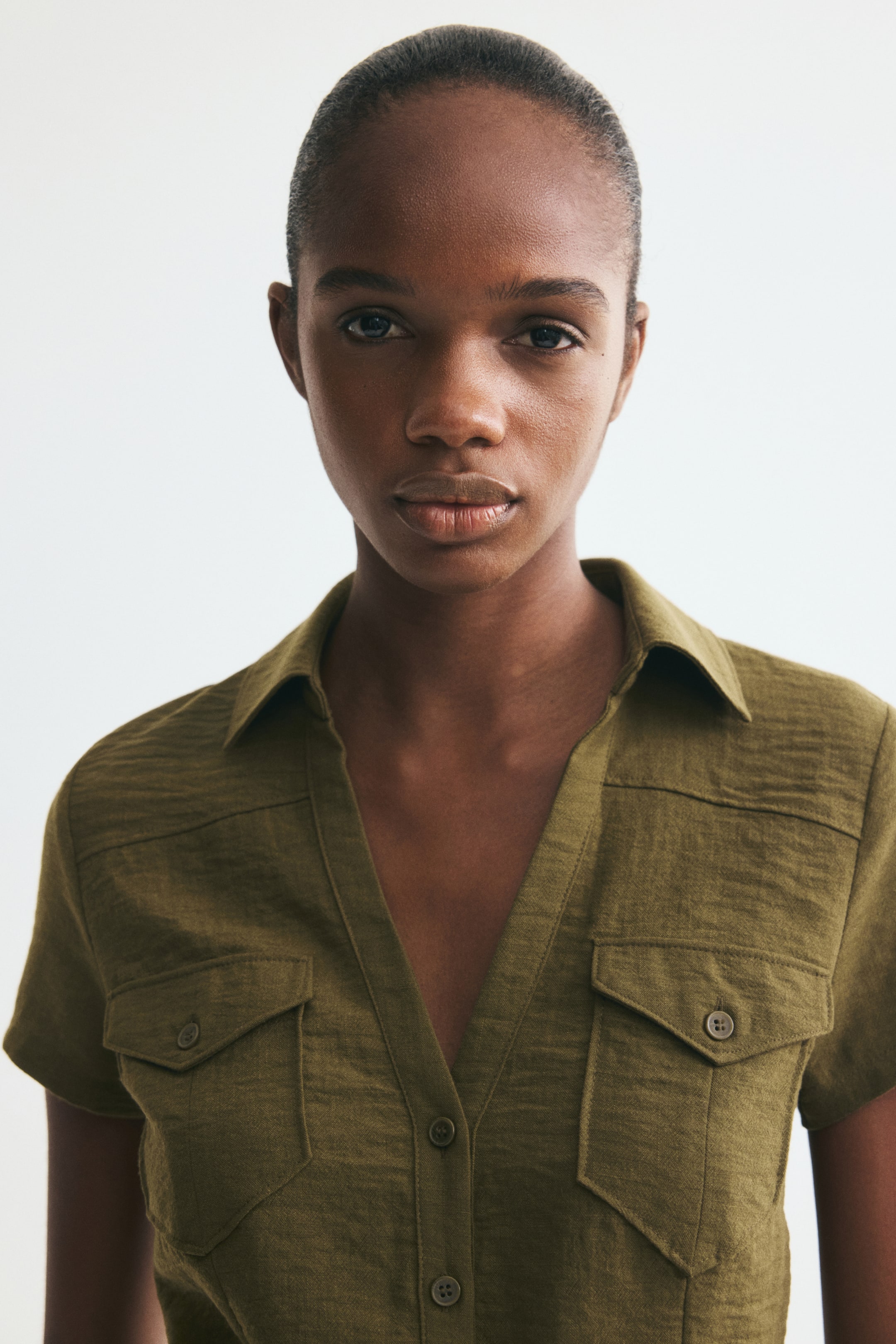 View larger image: The model is wearing a short-sleeved olive green shirt in woven fabric, featuring an open collar and front button fastening, with some buttons left undone. A yoke extends over the shoulders, and two chest flap pockets sit on the front.