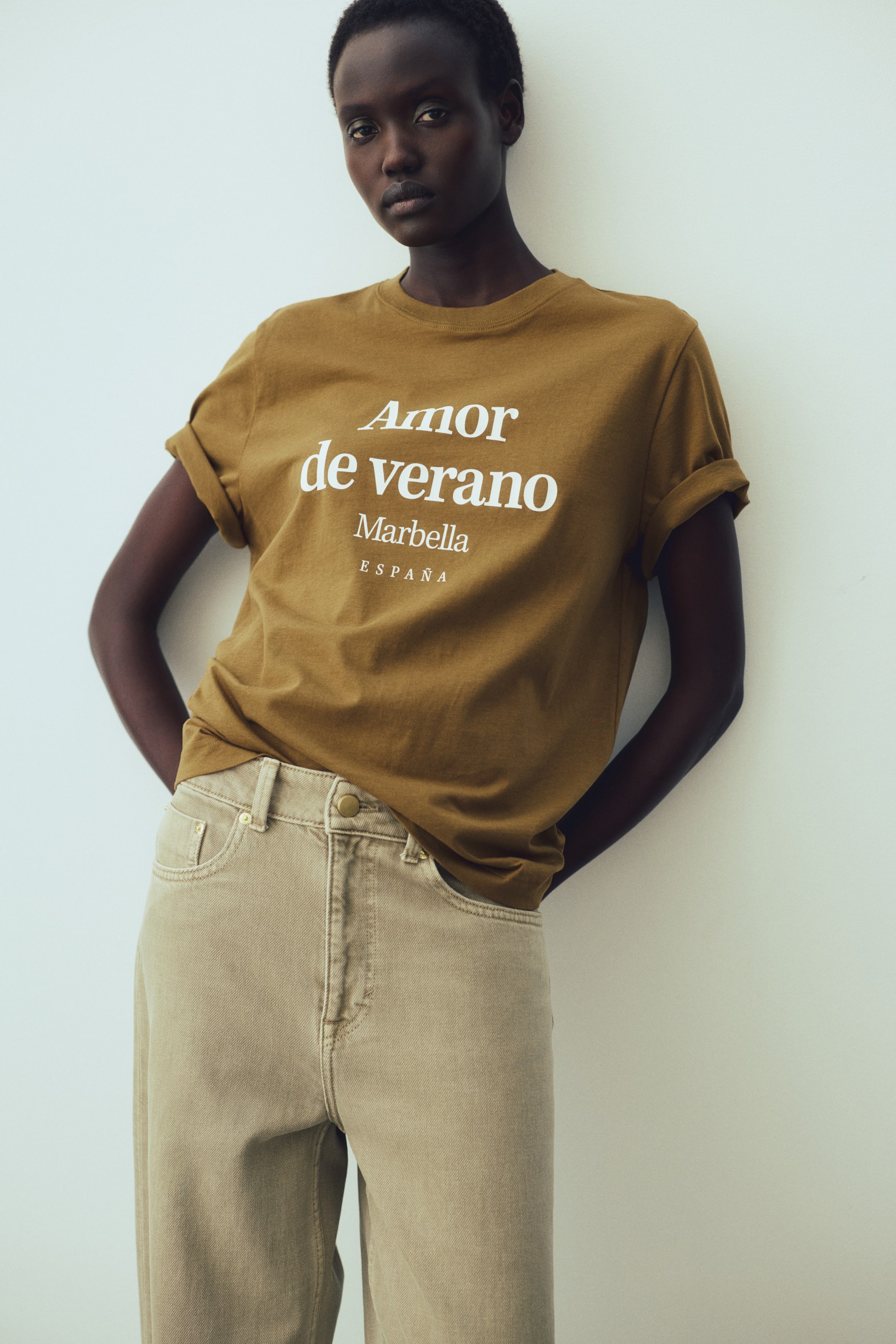 View larger image: An olive green T-shirt featuring a round neckline and white "Amor de verano Marbella ESPAÑA" text print is being worn by the model. Its short sleeves are cuffed, and the straight-cut hem is partially tucked into light beige trousers.