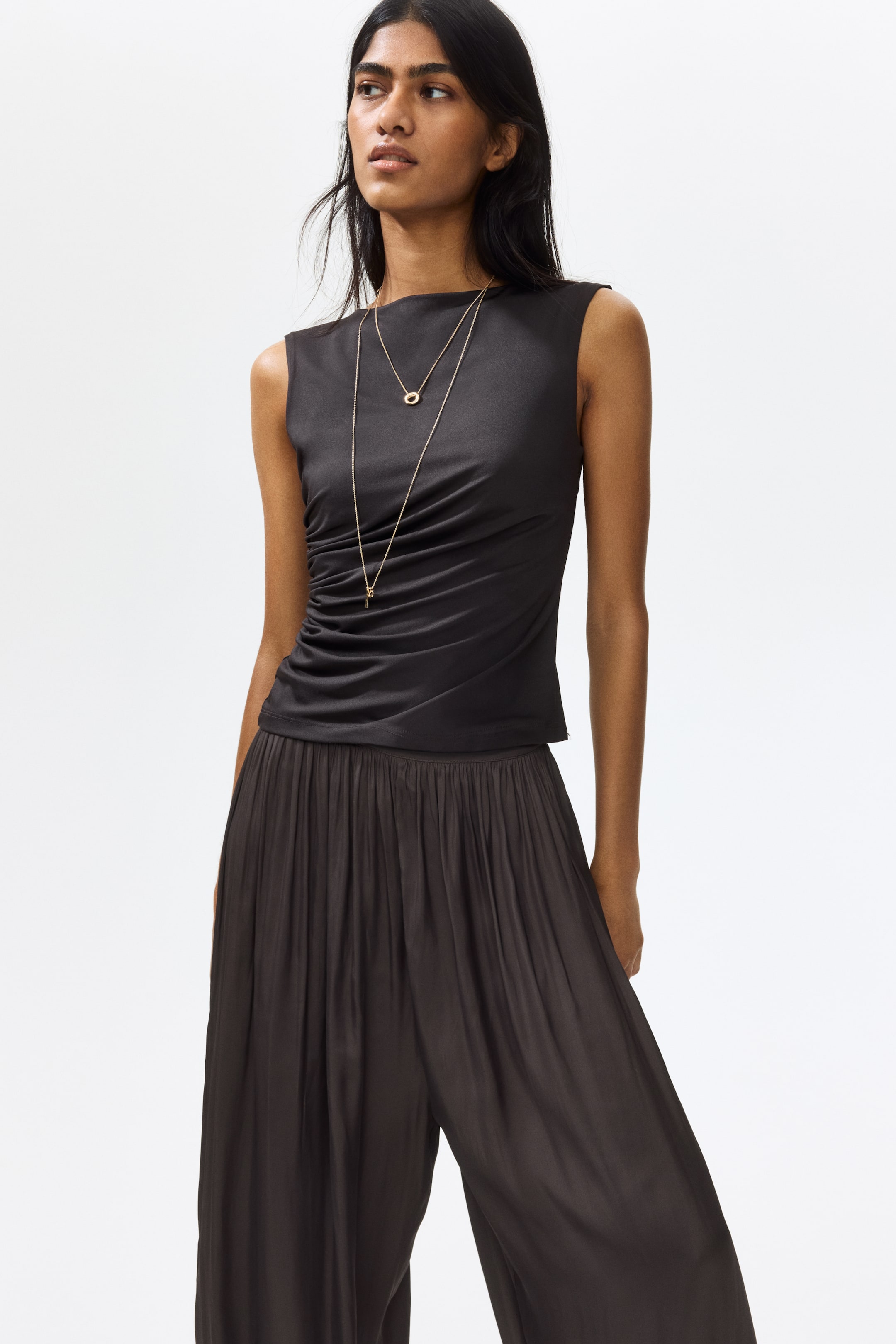 View larger image: The model is showcasing a fitted, sleeveless top crafted from jersey fabric with a subtle sheen. It boasts a boat neckline and a gathered seam on one side of the waist, giving rise to a softly draped effect. The ensemble is completed with dark, wide-leg pleated trousers and two layered gold necklaces.