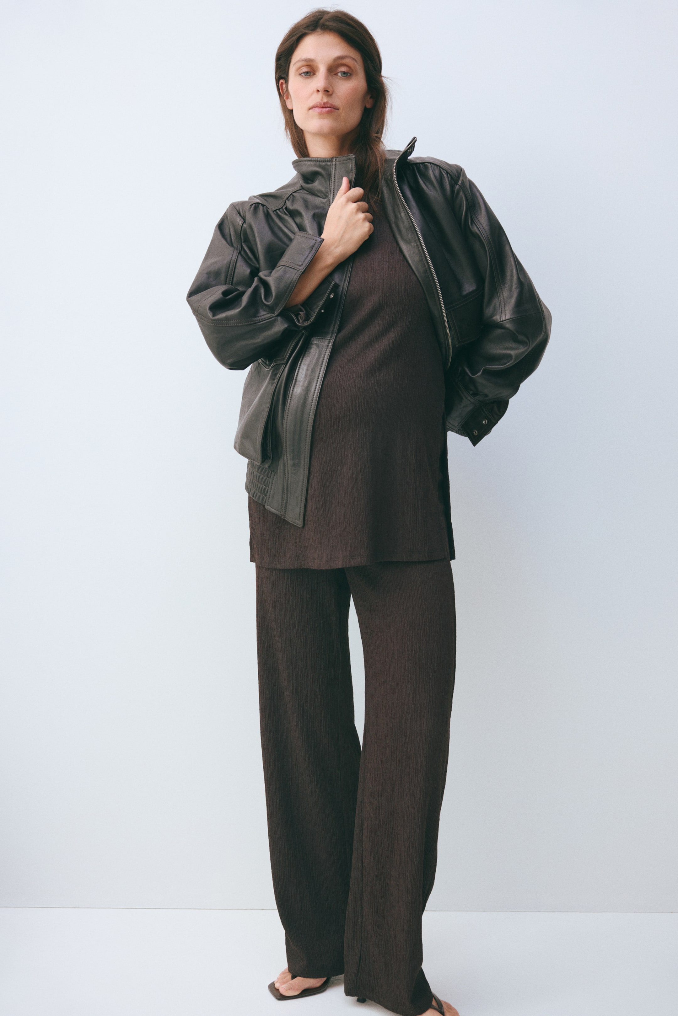 Prikaži veću sliku: Tamno braon rebrasti komplet od dva dela je predstavljen, koji sadrži tuniku širokog kroja i pantalone sa širokim nogavicama. Tamno braon kožna jakna sa uspravnim okovratnikom i naboranim ramenima nosi se otvoreno preko tunike.