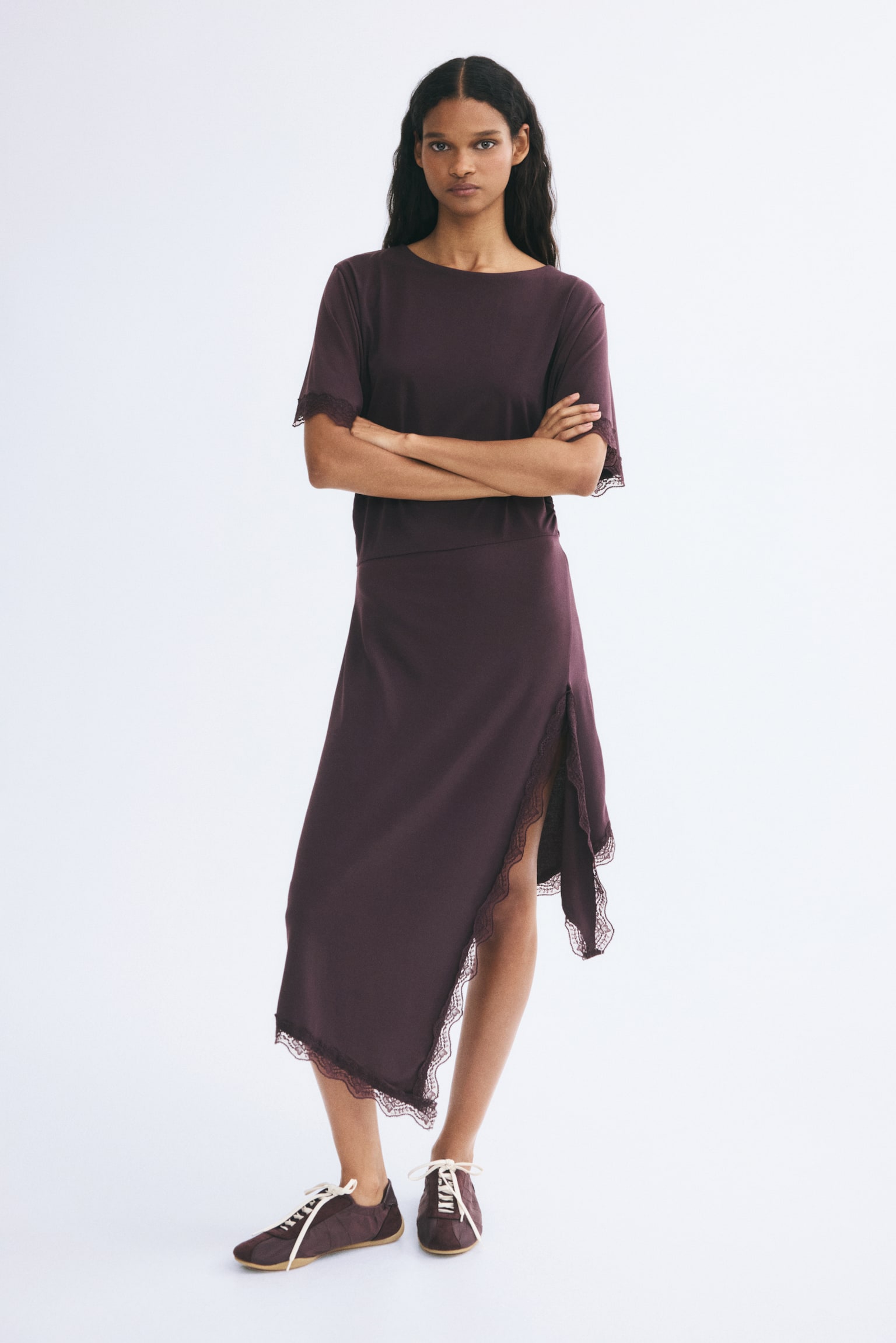 A dark plum, calf-length dress is worn by the model, featuring a round neckline, short sleeves with lace-trimmed cuffs, and gathers at the waist for a draped effect. The dress has an asymmetrical hem with a high side slit trimmed in matching lace, and is styled with coordinating plum runners. - 1