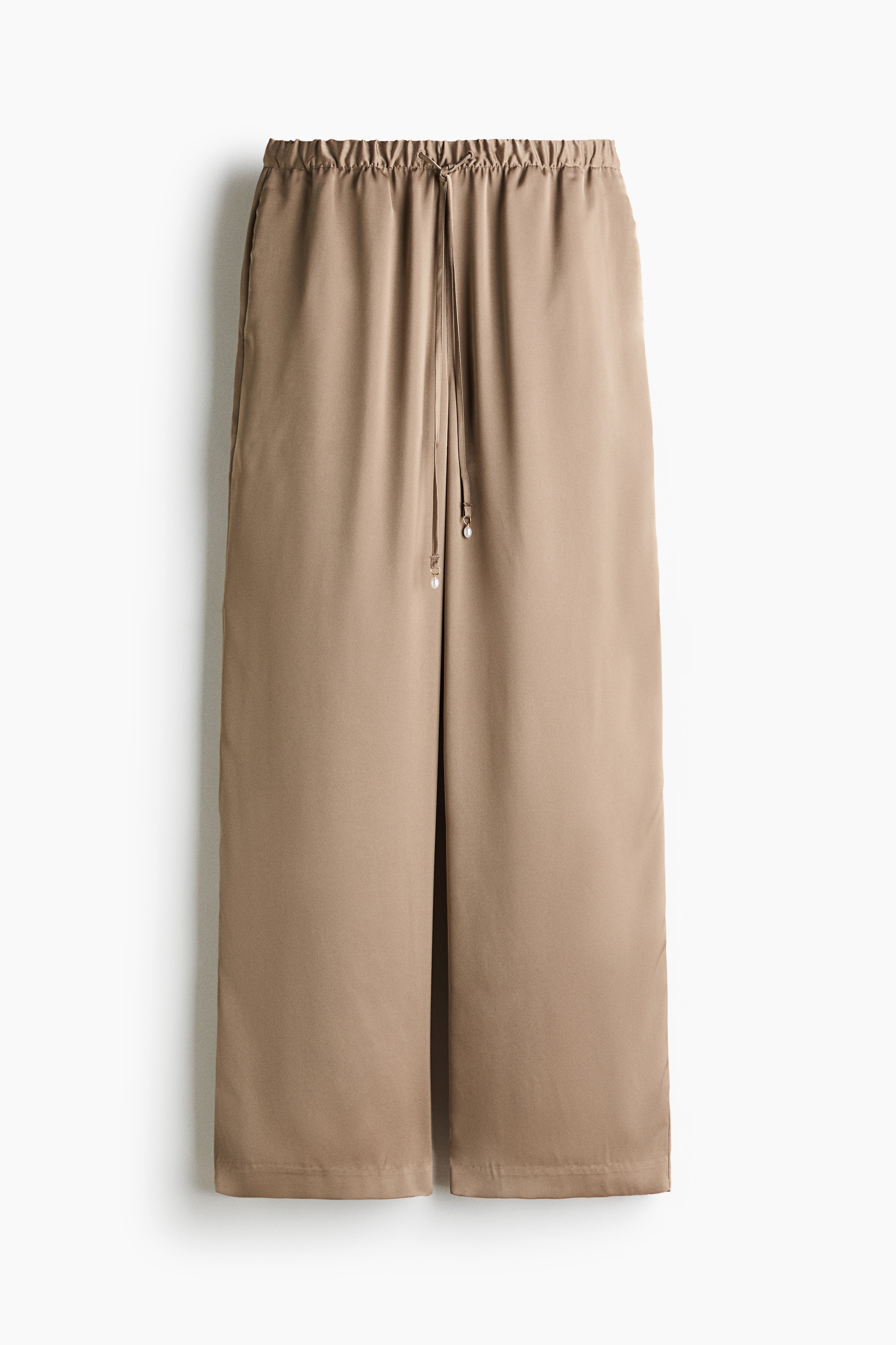 Wide-leg trousers in a light taupe hue are shown from the front, featuring a gathered elastic waistband with an adjustable matching drawstring. The slender drawstring cords hang down the centre, finished with small decorative pearl-like beads.