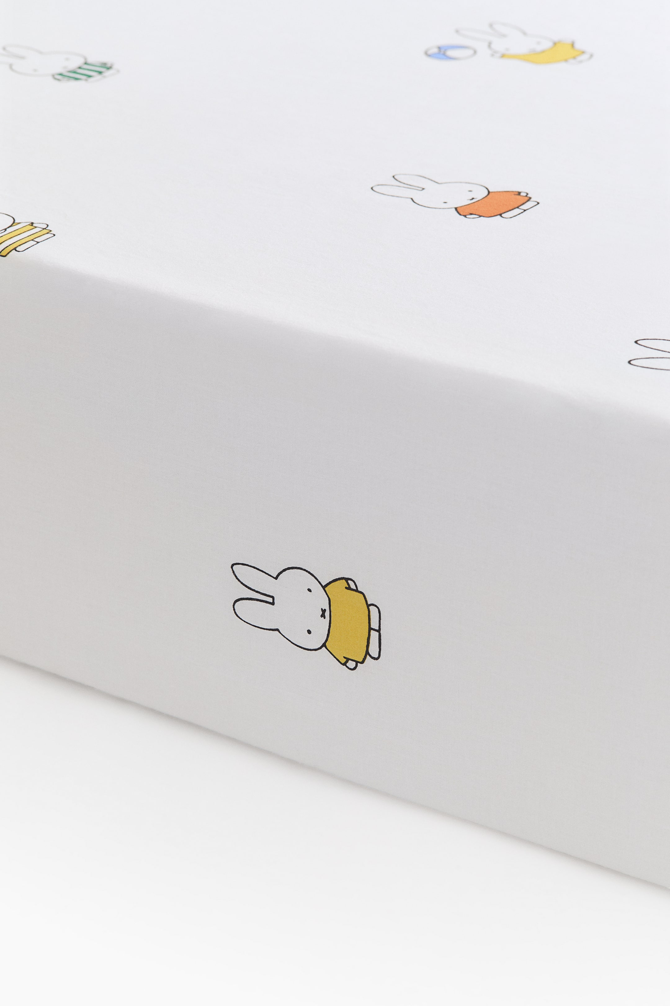 Zobraziť väčší obrázok: Biely textilný výrobok s opakujúcim sa vzorom ilustrácií králika Miffy, zobrazený z diagonálneho uhla. Ilustrácie ukazujú Miffyho oblečeného v žltom, oranžovom a pruhovanom zelenom oblečení.