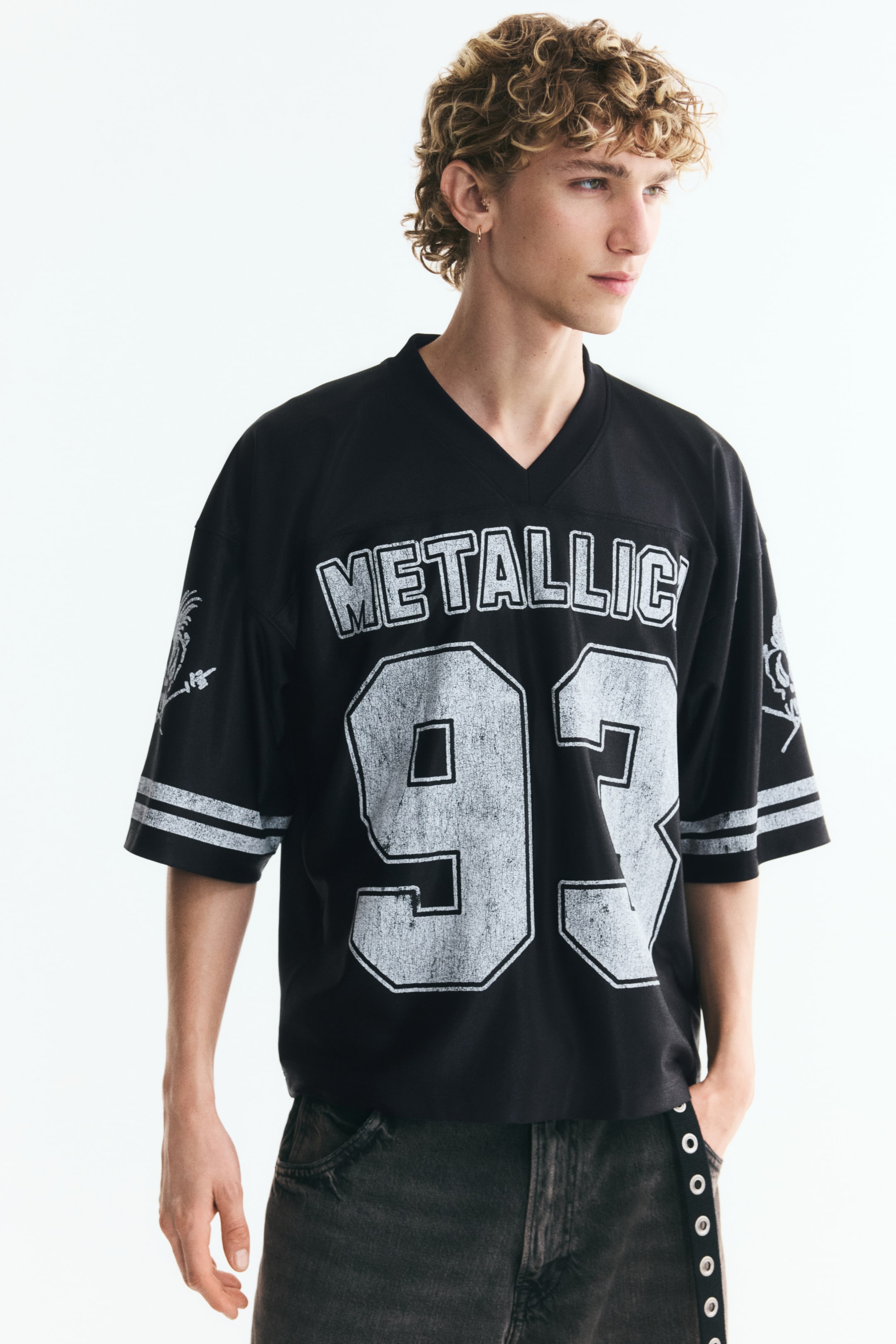 Büyük resmi görüntüle: Modelin üzerinde, ön kısmında baskılı "METALLICA 93" grafiği bulunan, fitilli V yaka, düşük omuzlu ve oversize kalıp siyah, jersey tarzı bir tişört yer alıyor. Bol kesimli tişört, yatay çizgili geniş kollara sahip olup koyu yıkamalı kot pantolonla kombinlenmiştir.