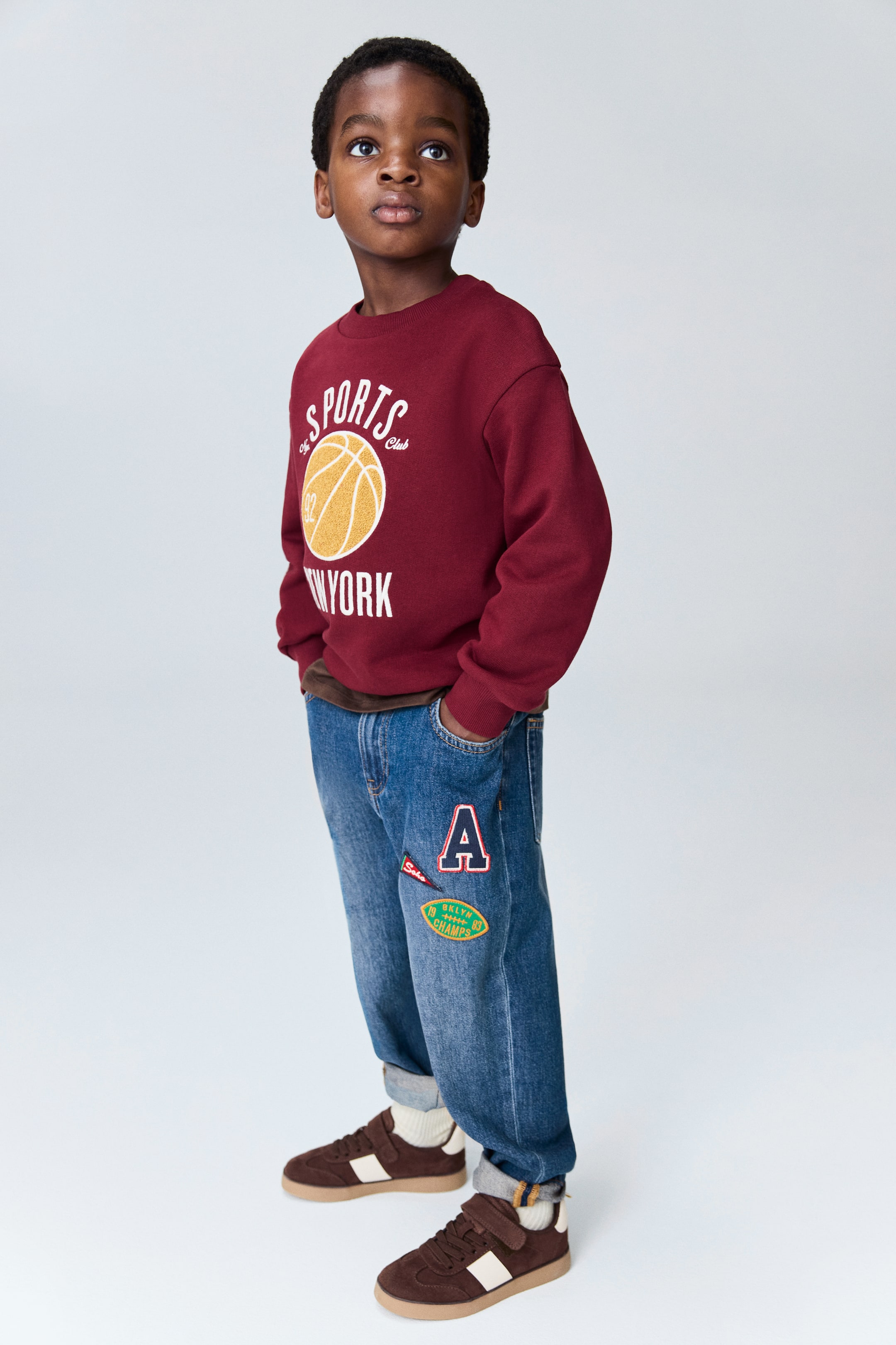View larger image: A maroon crew neck sweatshirt, featuring a "SPORTS Club NEW YORK" basketball graphic, is showcased with medium blue jeans adorned with multiple patches, including a red "A" and a "BKLYN CHAMPS" oval. White socks are visible above brown suede sneakers, which feature white accents and velcro straps, with the entire outfit being viewed from a 3/4 front angle.