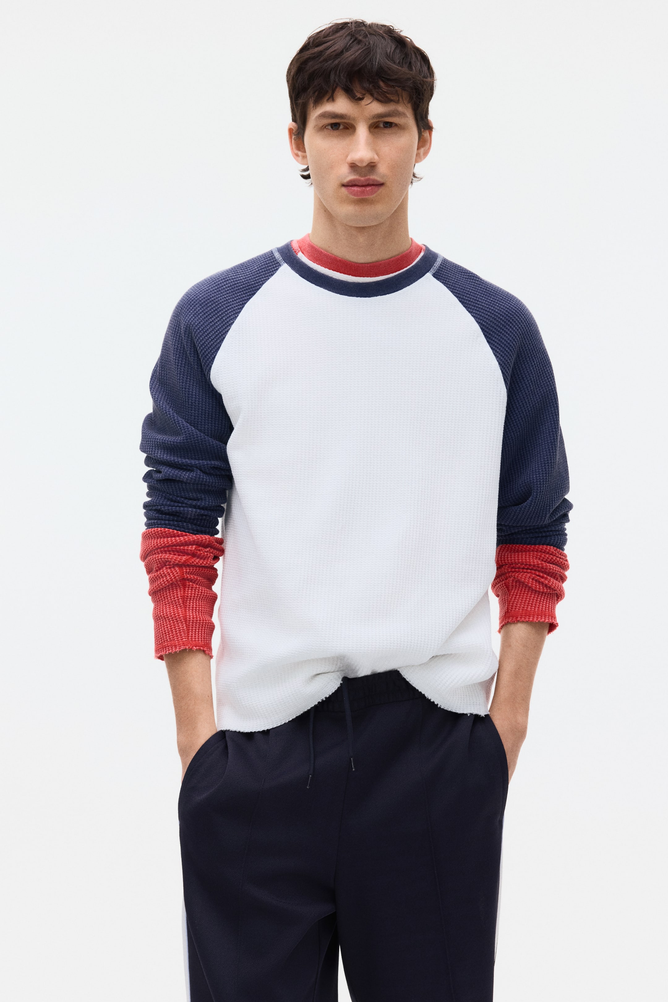 View larger image: A white and navy waffle-knit long-sleeve shirt is being worn, featuring a white body, navy raglan sleeves, and a raw-edged hem. A red long-sleeve shirt is visible underneath, appearing at the crew neck and with its ribbed cuffs extending past the main shirt's bunched navy sleeves.