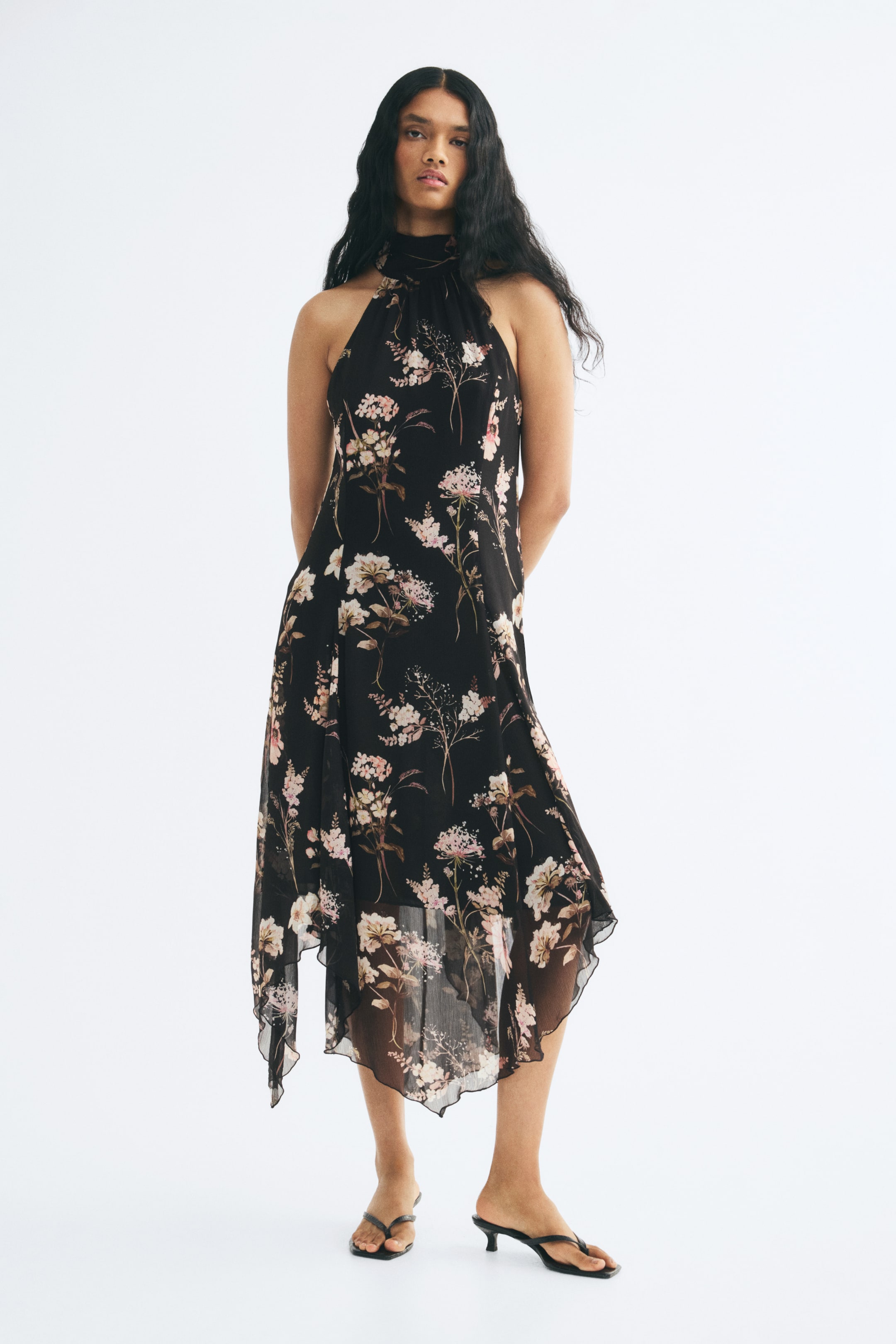 View larger image: The model is wearing a long black chiffon dress with a pink floral print, a gathered halter neckline, and an asymmetrical handkerchief hem. The dress is styled with black kitten-heel thong sandals.