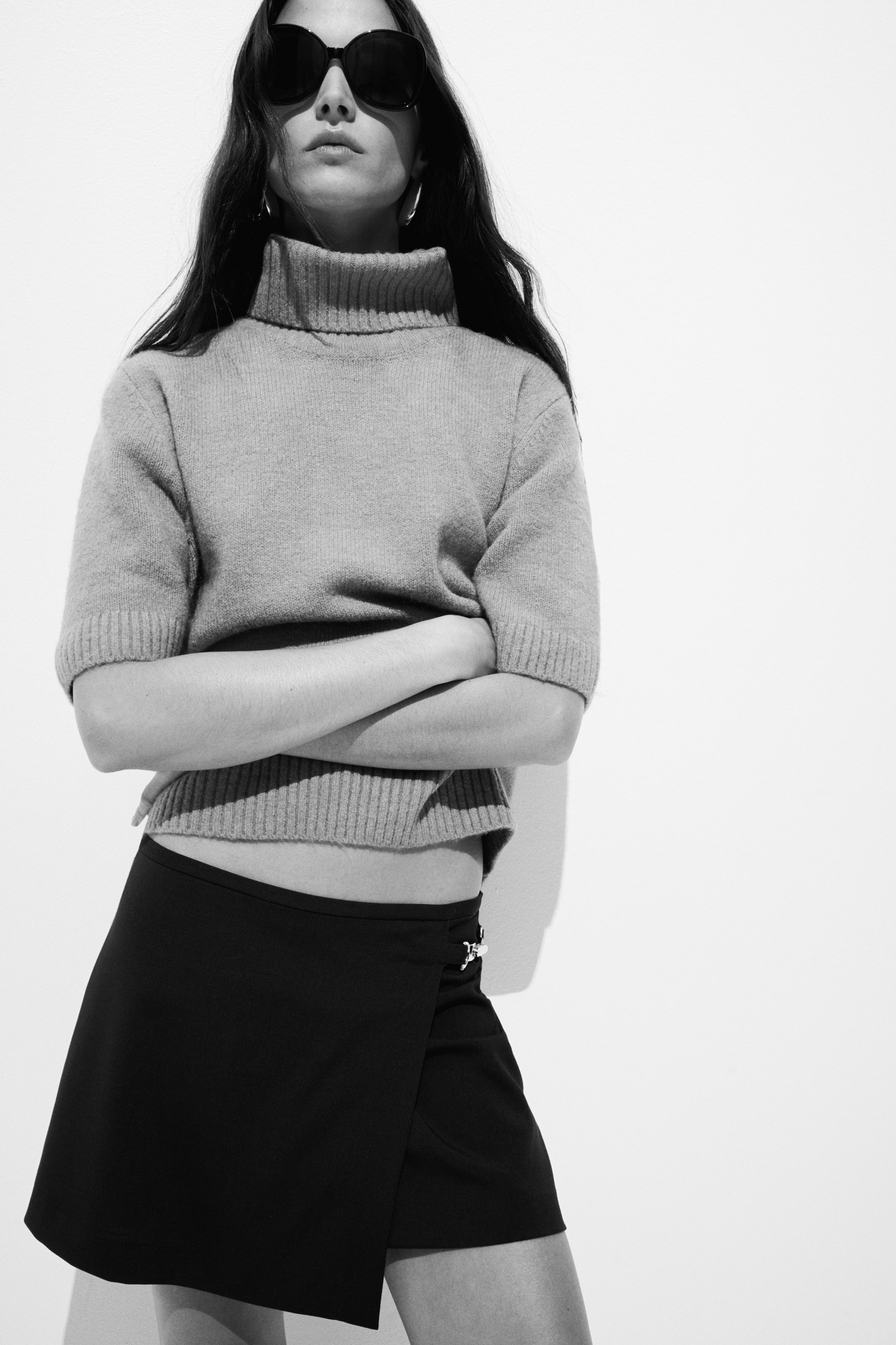 View larger image: Wearing a grey short-sleeved turtleneck jumper, showcasing a ribbed collar, ribbed cuffs, and a ribbed hem. Below the jumper, a black mini wrap skirt is worn, featuring a silver-toned buckle at the waist.