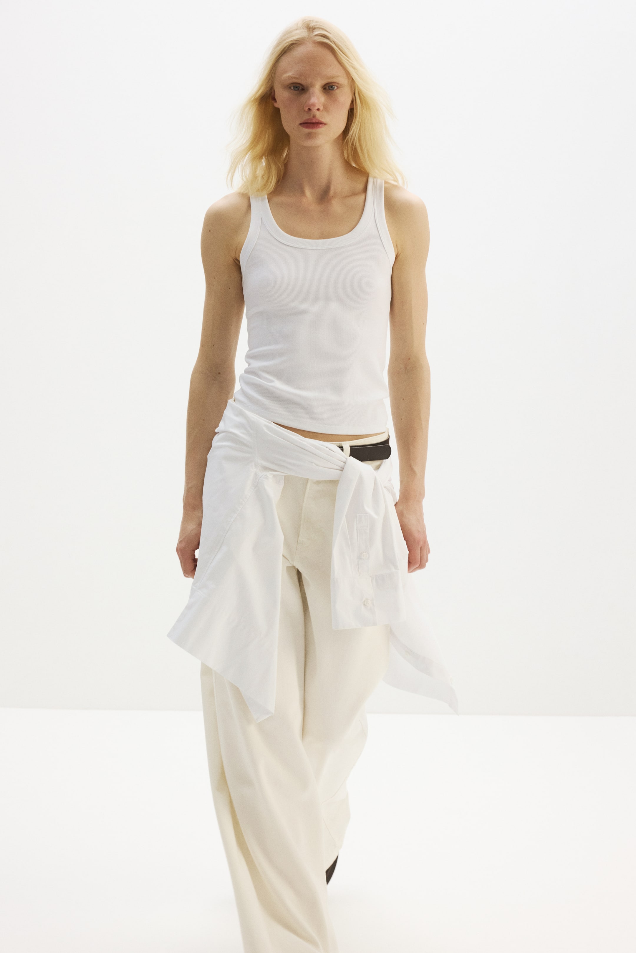 Größeres Bild ansehen: Ein eng anliegendes weißes geripptes Tanktop mit Rundhalsausschnitt, tiefen Armlöchern und einem geraden Saum wird von dem Model getragen. Ein weißes Hemd ist um die Taille des Models gebunden und kombiniert das Tanktop mit cremefarbenen, weit geschnittenen Hosen.
