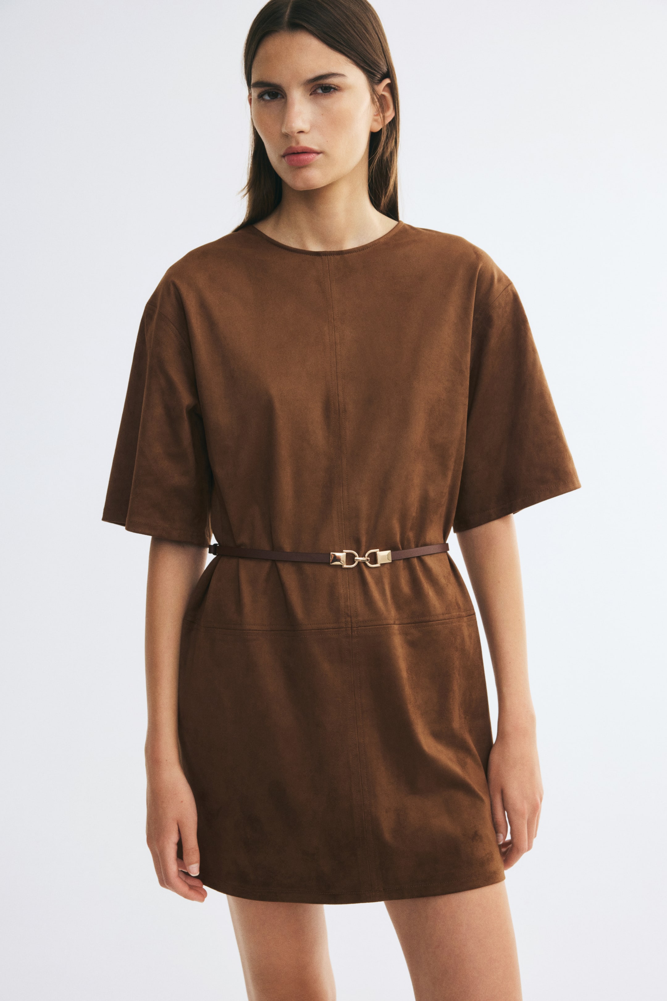 Voir une image plus grande: La mannequin porte une robe courte brune en tissu duveteux, dotée d’un col rond, d’épaules tombantes et de manches courtes amples. La robe est cintrée à la taille à l’aide d’une fine ceinture brune munie d’une boucle de ton doré, mettant en valeur les coutures visibles sur le devant.