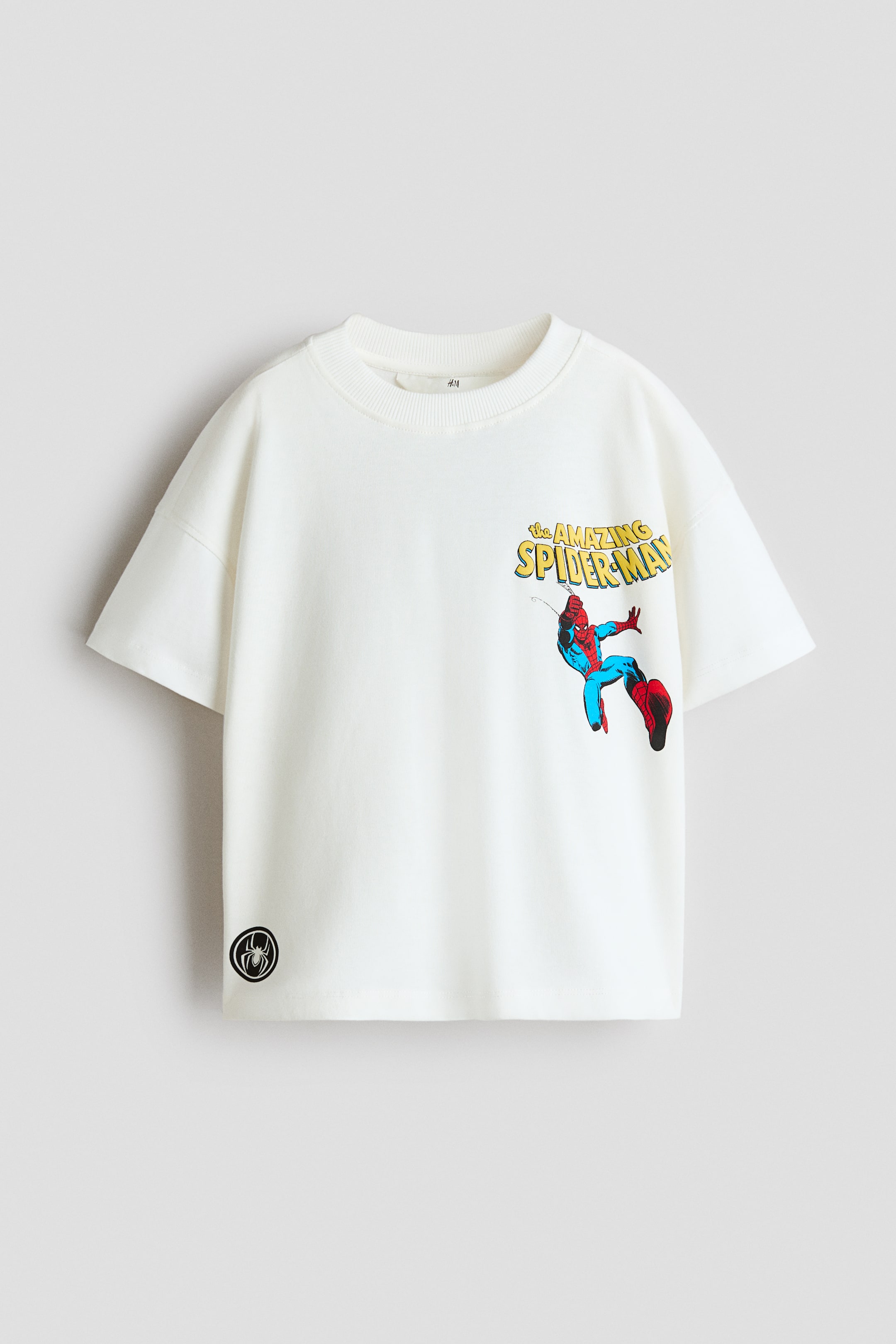 View larger image: An oversized off-white cotton jersey T-shirt is shown from the front, featuring a round, rib-trimmed neckline and dropped shoulders. The T-shirt features a graphic of Spider-Man swinging beneath the yellow text “the AMAZING SPIDER-MAN” on the left chest, along with a small circular black spider patch on the lower right hem.