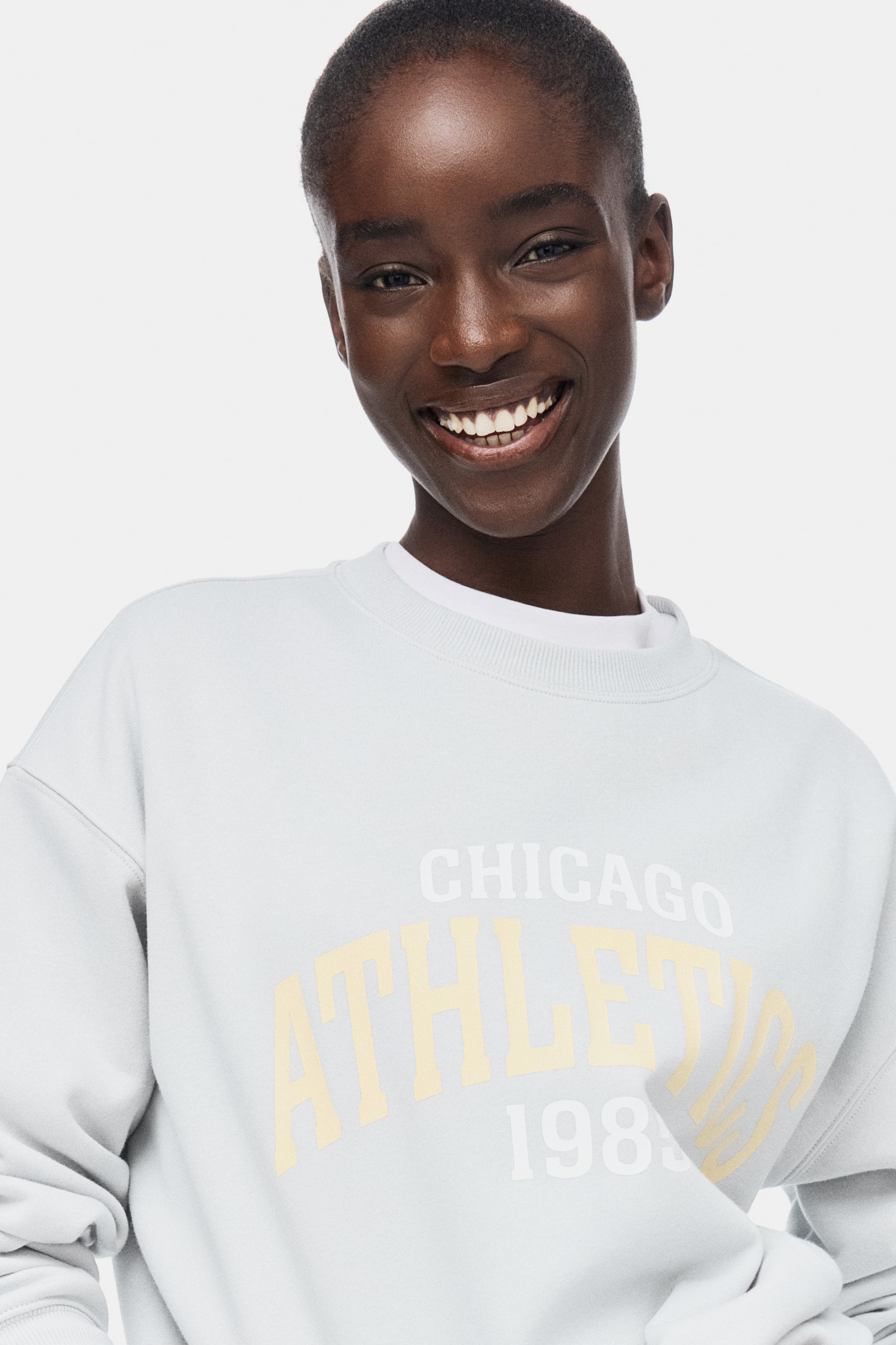View larger image: A light blue crew neck sweatshirt showcases the text "CHICAGO ATHLETICS 198" across its front, with "CHICAGO" in white, "ATHLETICS" in light yellow, and "198" in white. It is stylishly layered over a white crew neck t-shirt, visible at the neckline.