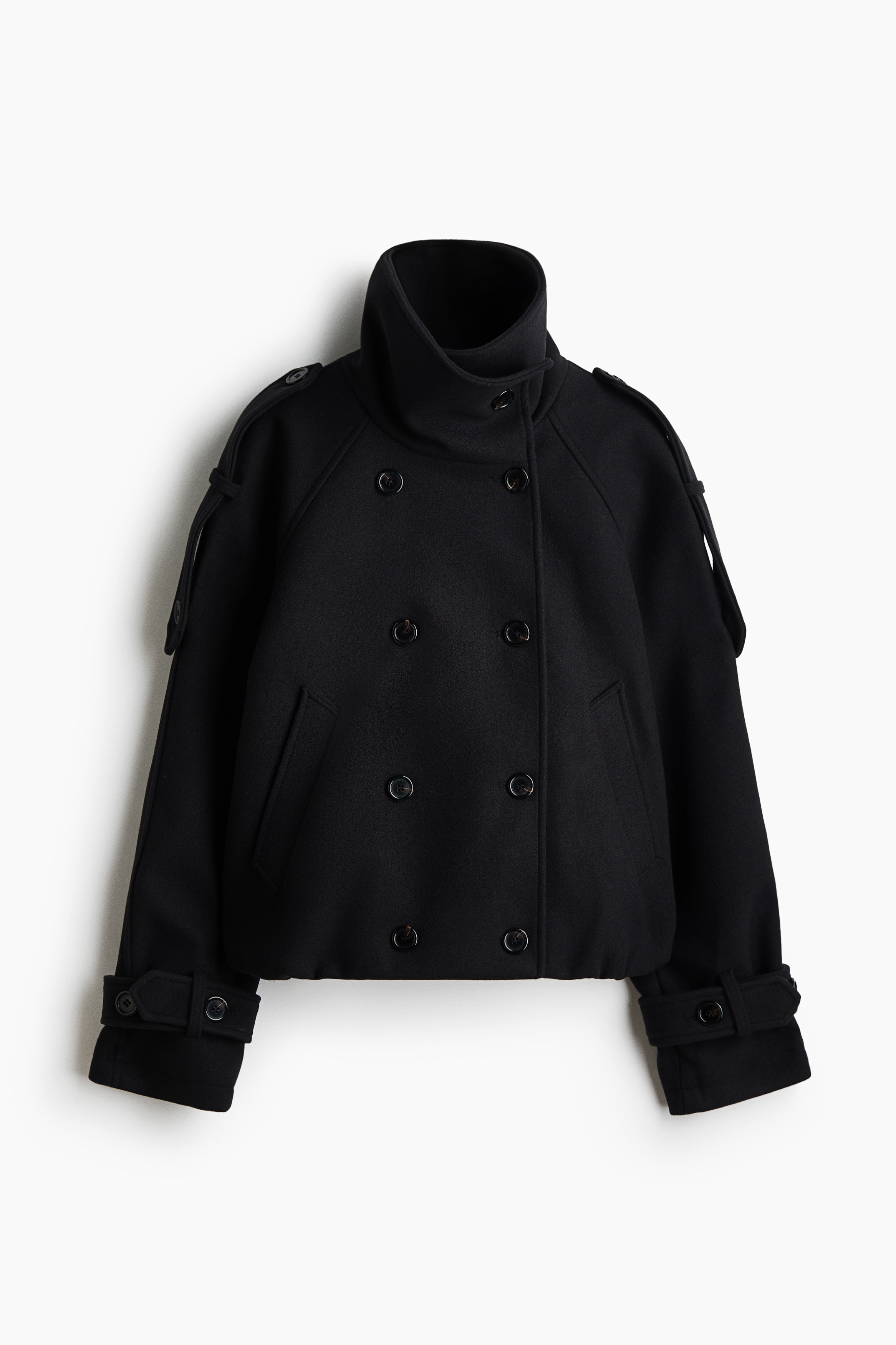 A black, double-breasted jacket in felted fabric is being worn by the model over a black roll-neck top. The oversized jacket features a wide collar, buttoned shoulder tabs, and welt front pockets, with the model placing one hand inside the pocket.