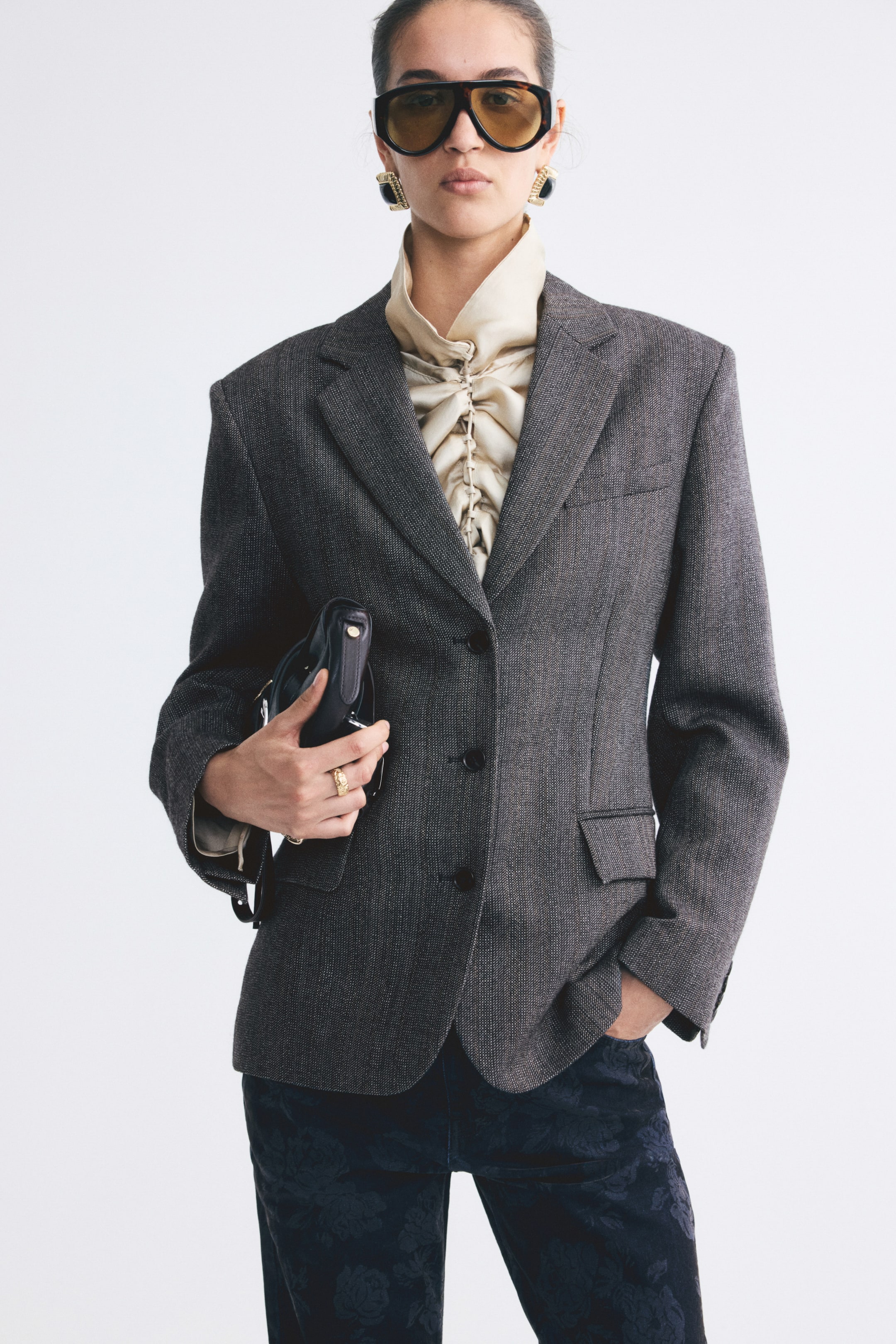 View larger image: The model is wearing a single-breasted blazer in a textured grey wool weave, layered over a ruched cream blouse. The blazer features structured shoulders, notch lapels, front button fastening, and jetted flap pockets, with the model carrying a small black clutch bag.