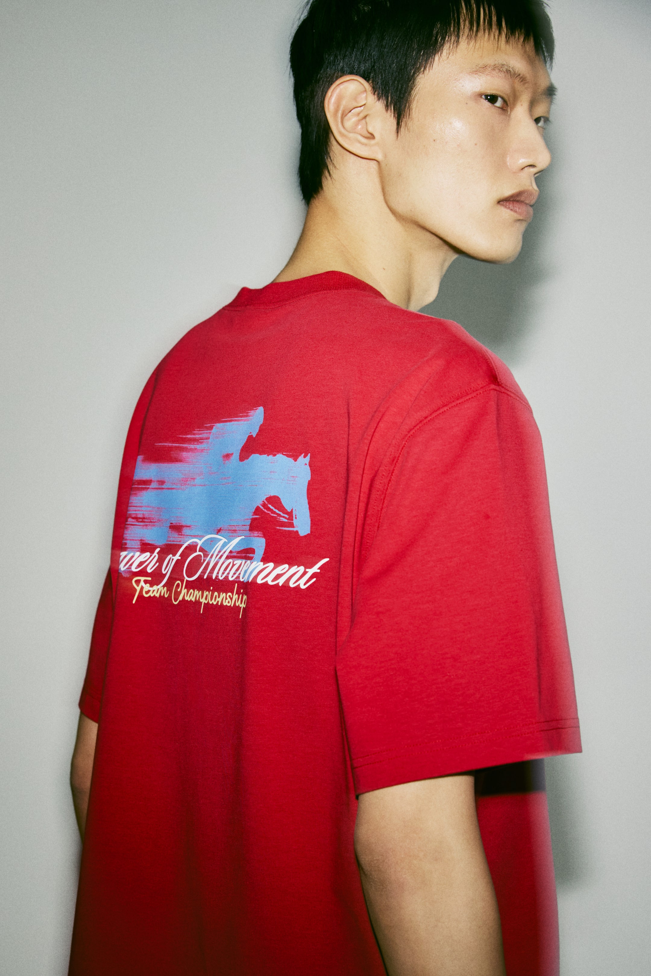 Xem ảnh lớn hơn: Một chiếc áo phông màu đỏ có in một họa tiết ở mặt sau, nổi bật hình bóng màu xanh nhạt của một người cưỡi ngựa kèm các đường ngang tạo hiệu ứng chuyển động. Phía dưới hình bóng, dòng chữ "Power of Movement Team Championship" xuất hiện bằng phông chữ script và sans-serif màu trắng.