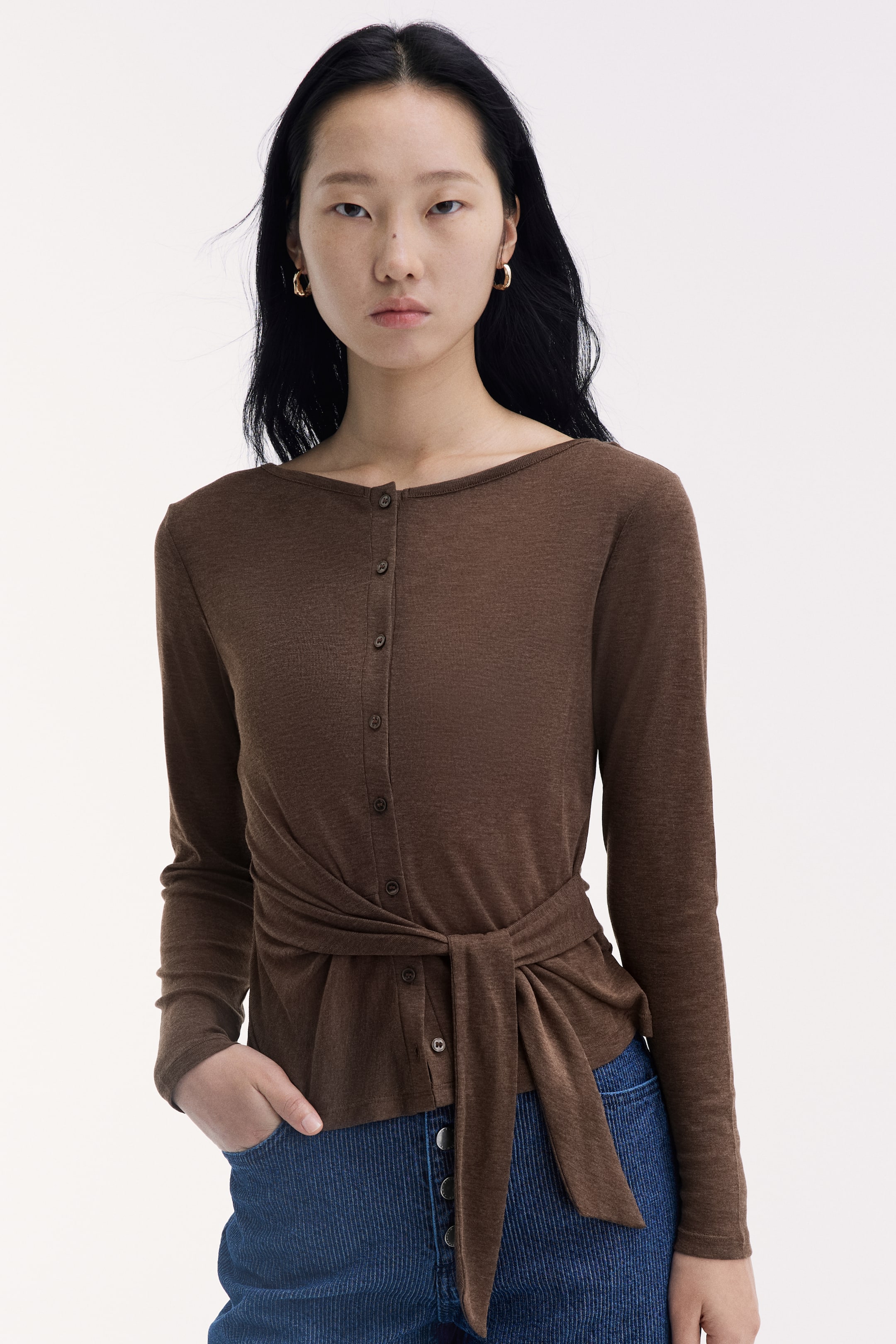 View larger image: A brown, long-sleeved knit top with a boat neck and a button-down front is being worn by the model. The top features a fabric panel wrapped and tied at the waist, and is paired with blue textured trousers.