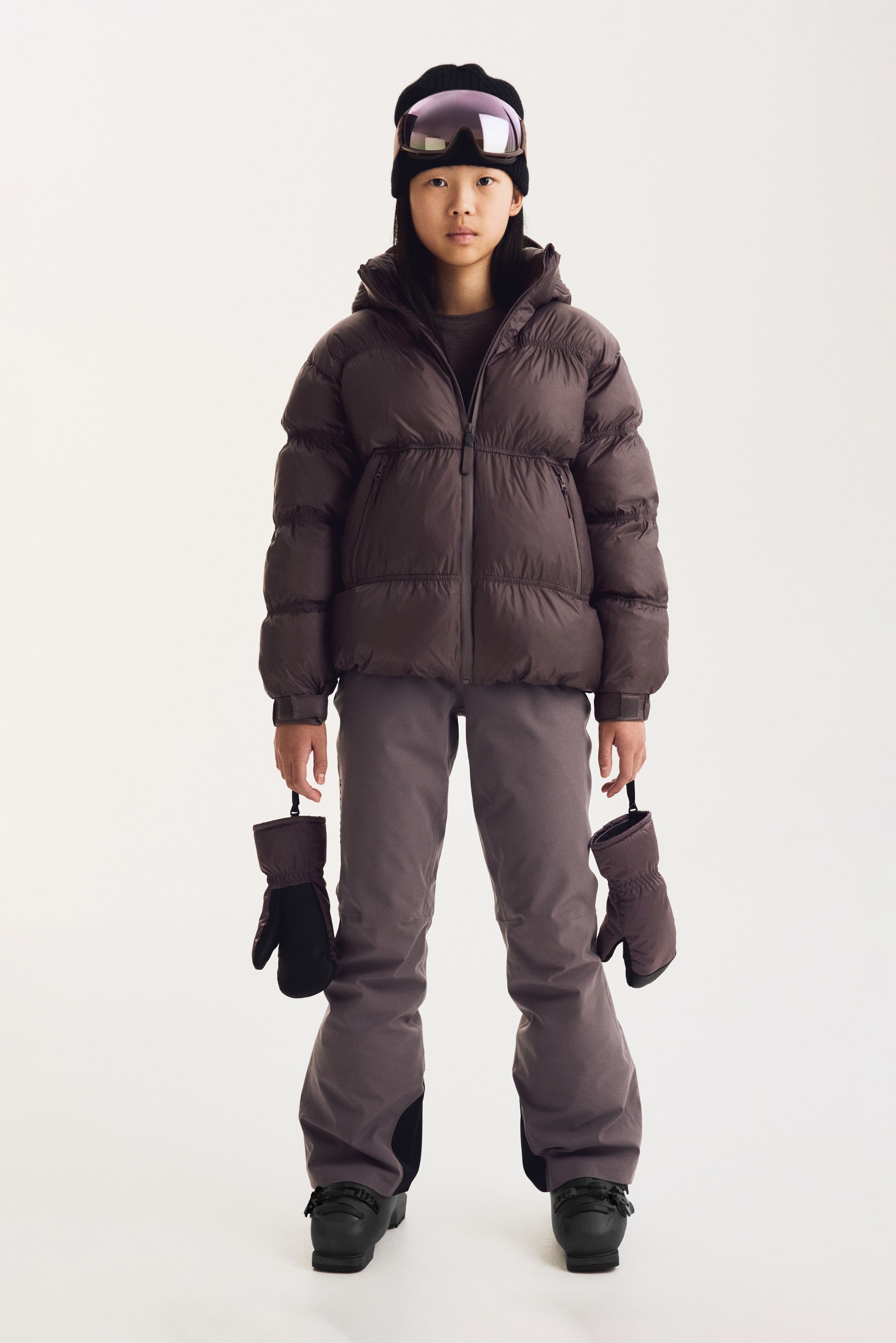 Une doudoune violet poussiÃ©reux est prÃ©sentÃ©e de face, mettant en valeur un design Ã  capuche avec fermeture zippÃ©e centrale et un matelassage horizontal froncÃ© qui crÃ©e un effet plissÃ©. La veste prÃ©sente deux poches latÃ©rales inclinÃ©es zippÃ©es et des poignets rÃ©glables avec bandes auto-agrippantes.