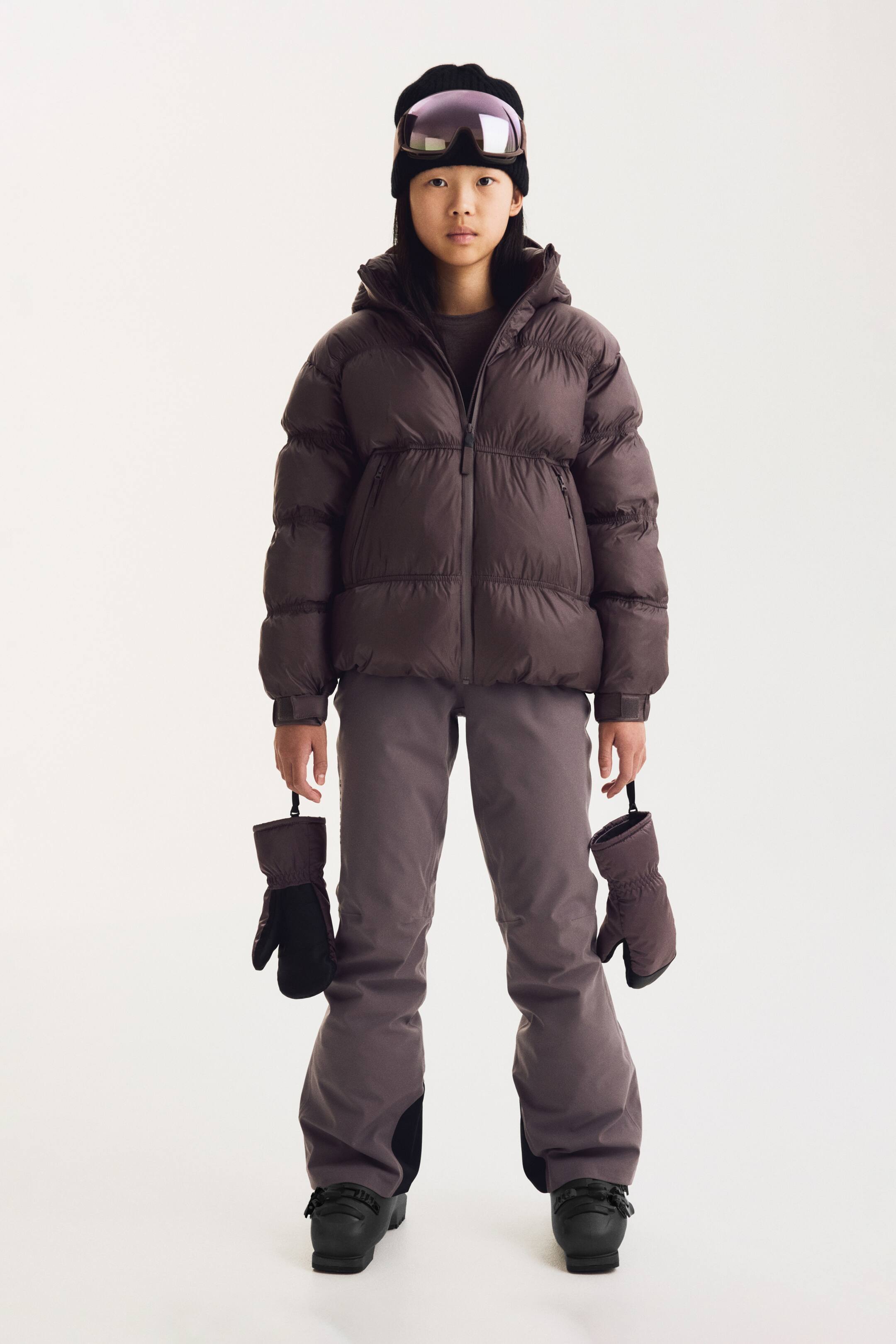 Agrandir l'image: Un mannequin porte une doudoune à capuche marron foncé présentant un matelassage horizontal et une fermeture zippée intégrale, assortie d’un pantalon de neige gris-brun, de face. Le mannequin tient des moufles assorties et porte un bonnet noir avec des lunettes de ski ainsi que des bottes de ski noires.