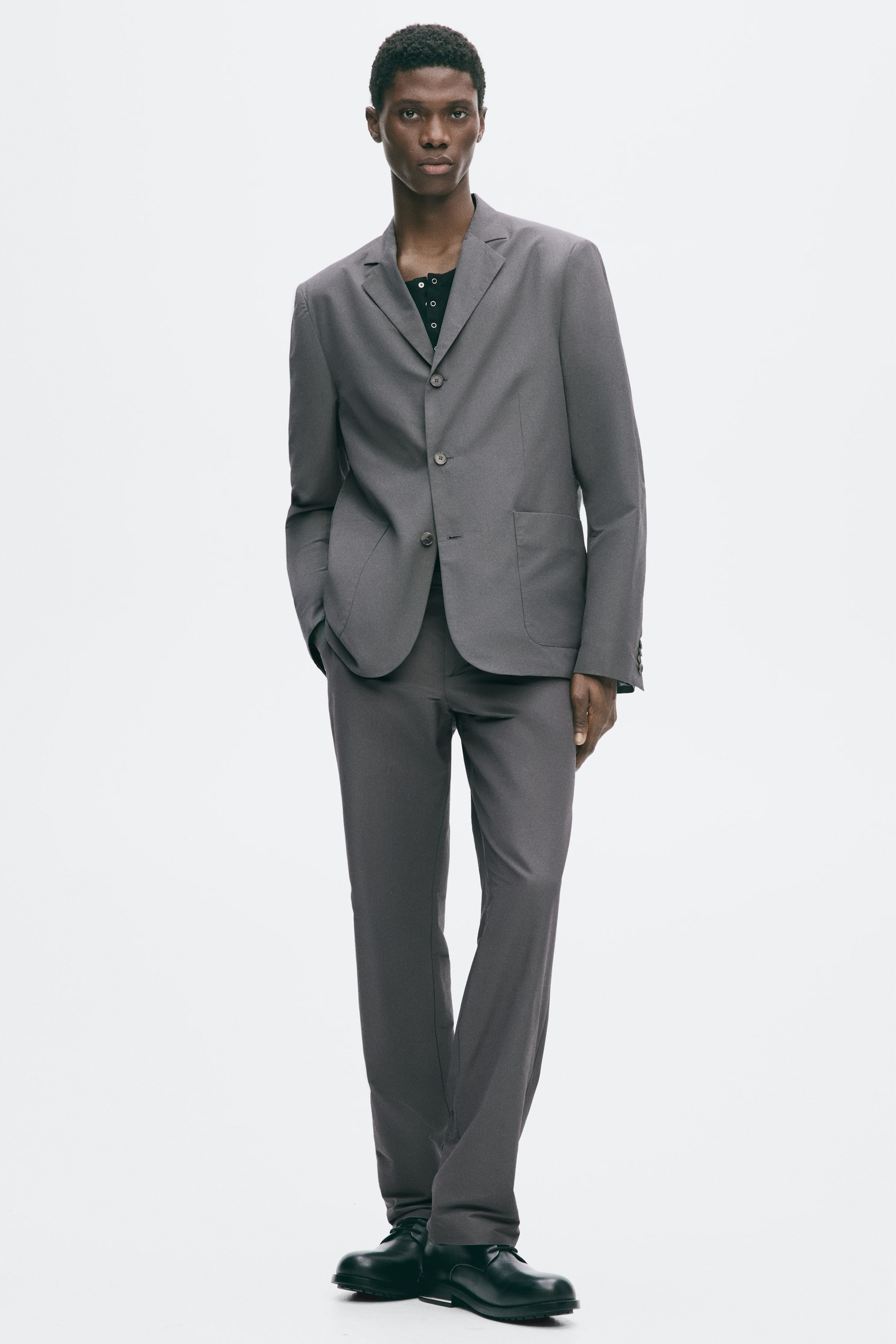 View larger image: The model is wearing a gray single-breasted jacket featuring notch lapels, front button closures, and patch pockets. The relaxed-fit jacket is styled with matching trousers and a dark, buttoned underlayer.