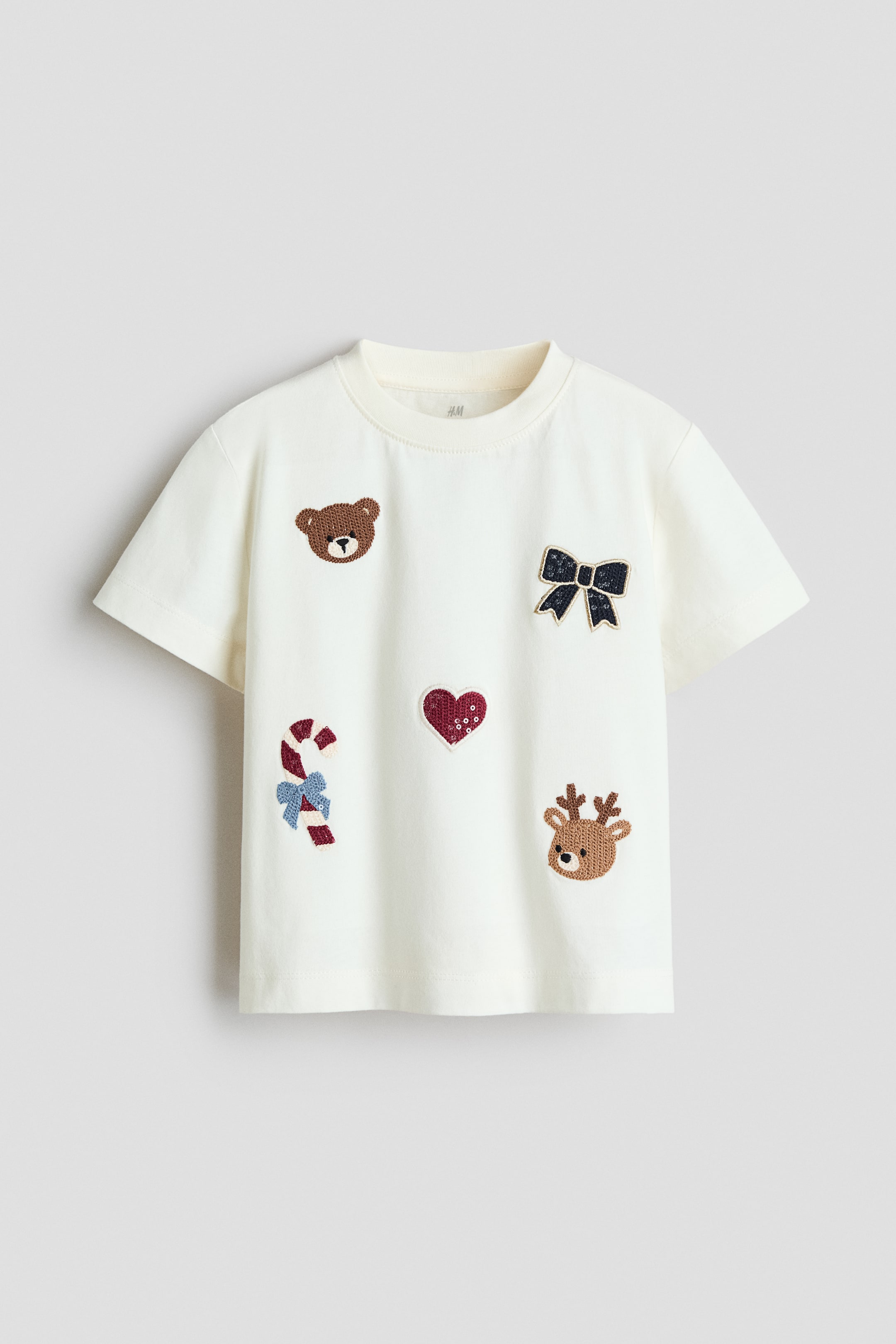 View larger image: A cream-colored, short-sleeved T-shirt is shown from the front, featuring a ribbed crewneck. The front of the shirt is decorated with five distinct patches, including a teddy bear head, a black bow, a red sequined heart, a candy cane, and a reindeer head.