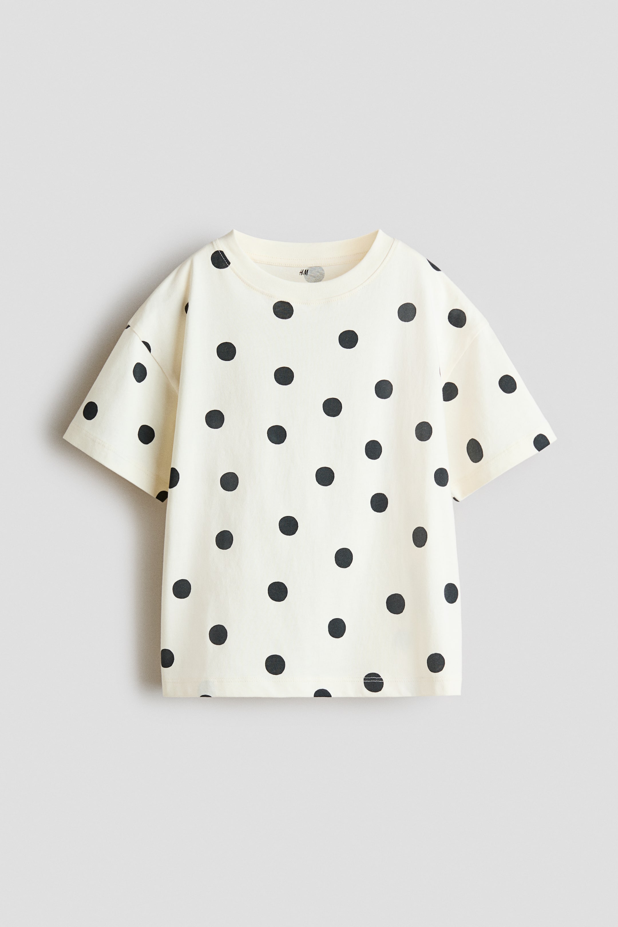Agrandir l'image: Un T-shirt à coupe décontractée en jersey de coton imprimé avec un col rond bordé de côtes et des épaules tombantes se présente de face, affichant une base blanc cassé avec un motif à pois noirs sur l'ensemble.