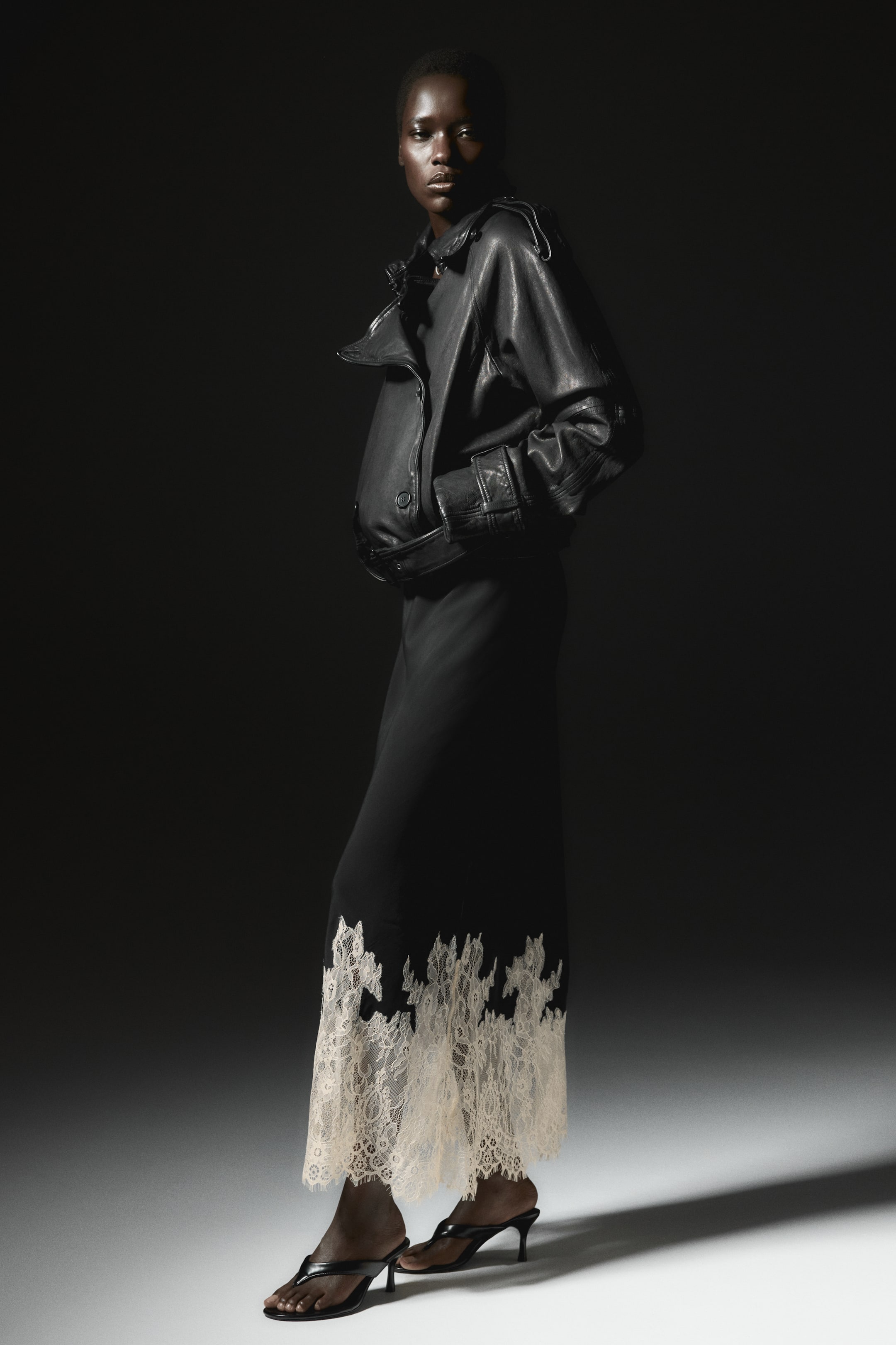 View larger image: The model is wearing a long, fitted black skirt featuring a wide, cream lace trim at the hem. It's styled with a black leather jacket and black heeled sandals.