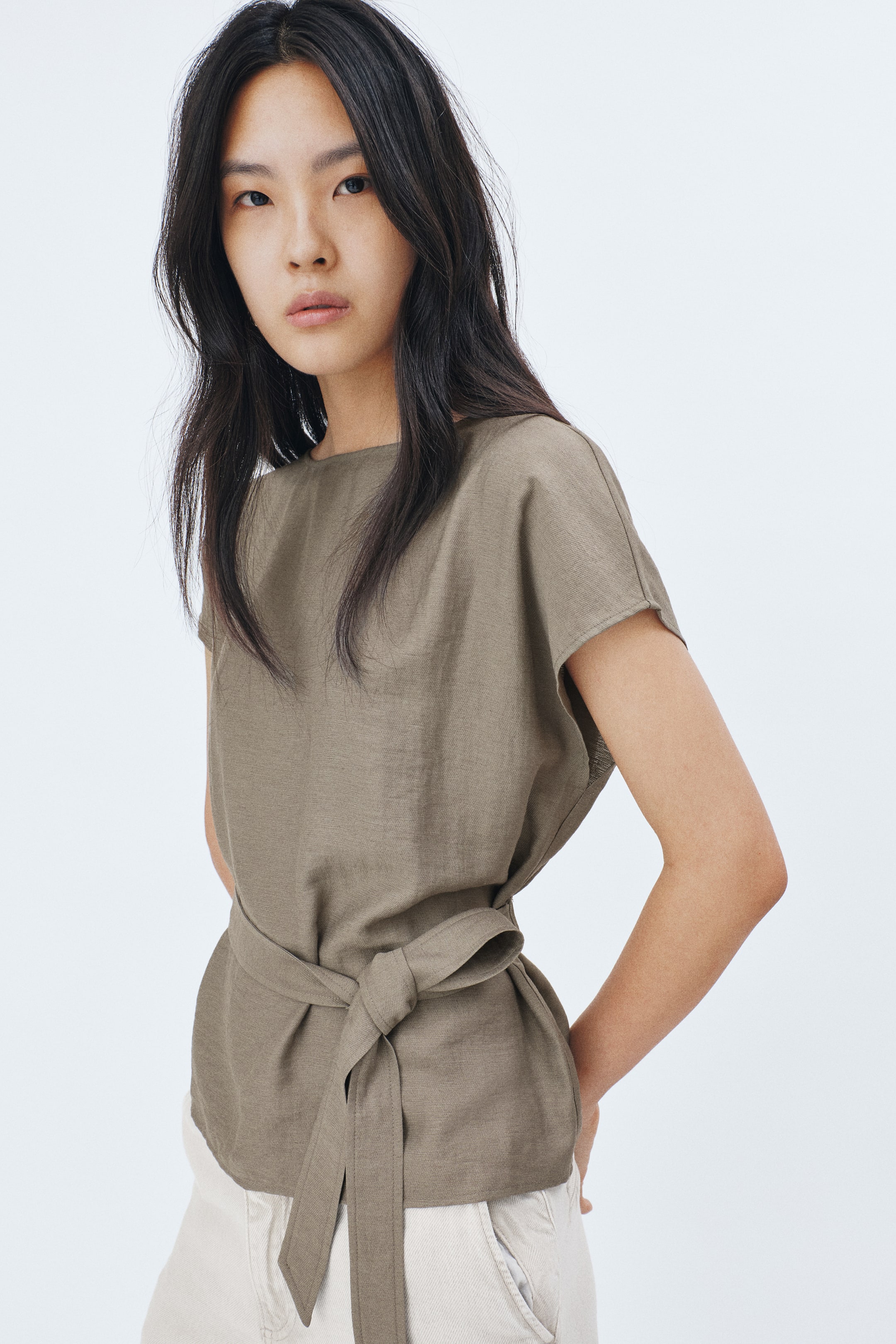 View larger image: Model tersebut memakai baju khaki yang diperbuat daripada campuran viscose dengan sedikit linen, menampilkan leher bot dan lengan cap. Tali pinggang lebar mengikat pinggang dan diikat di bahagian depan.