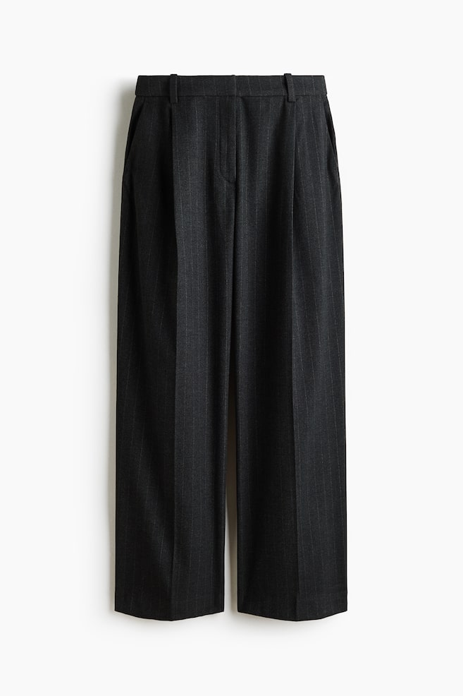 Se muestra un par de pantalones holgados de rayas diplomáticas en color gris oscuro, vistos de frente, con cintura alta y trabillas para cinturón, asà como pliegues frontales. El diseño incluye bolsillos laterales diagonales y marcadas lÃneas de pliegue que recorren el centro de cada pierna ancha. - 1