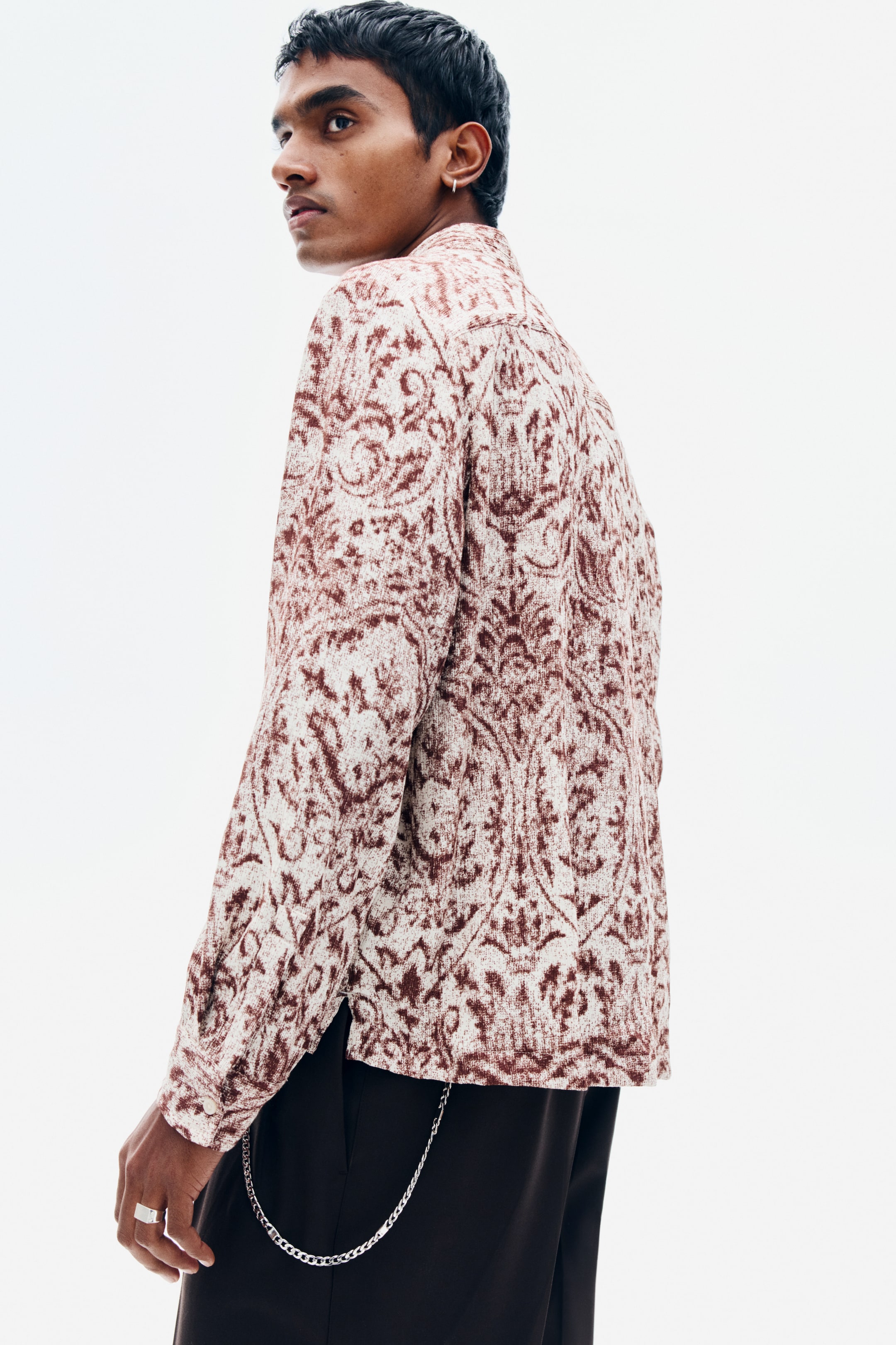 View larger image: The model is wearing a long-sleeved shirt featuring an all-over brown ornamental pattern on a cream-coloured soft weave. The shirt is styled with dark trousers featuring a silver pocket chain.