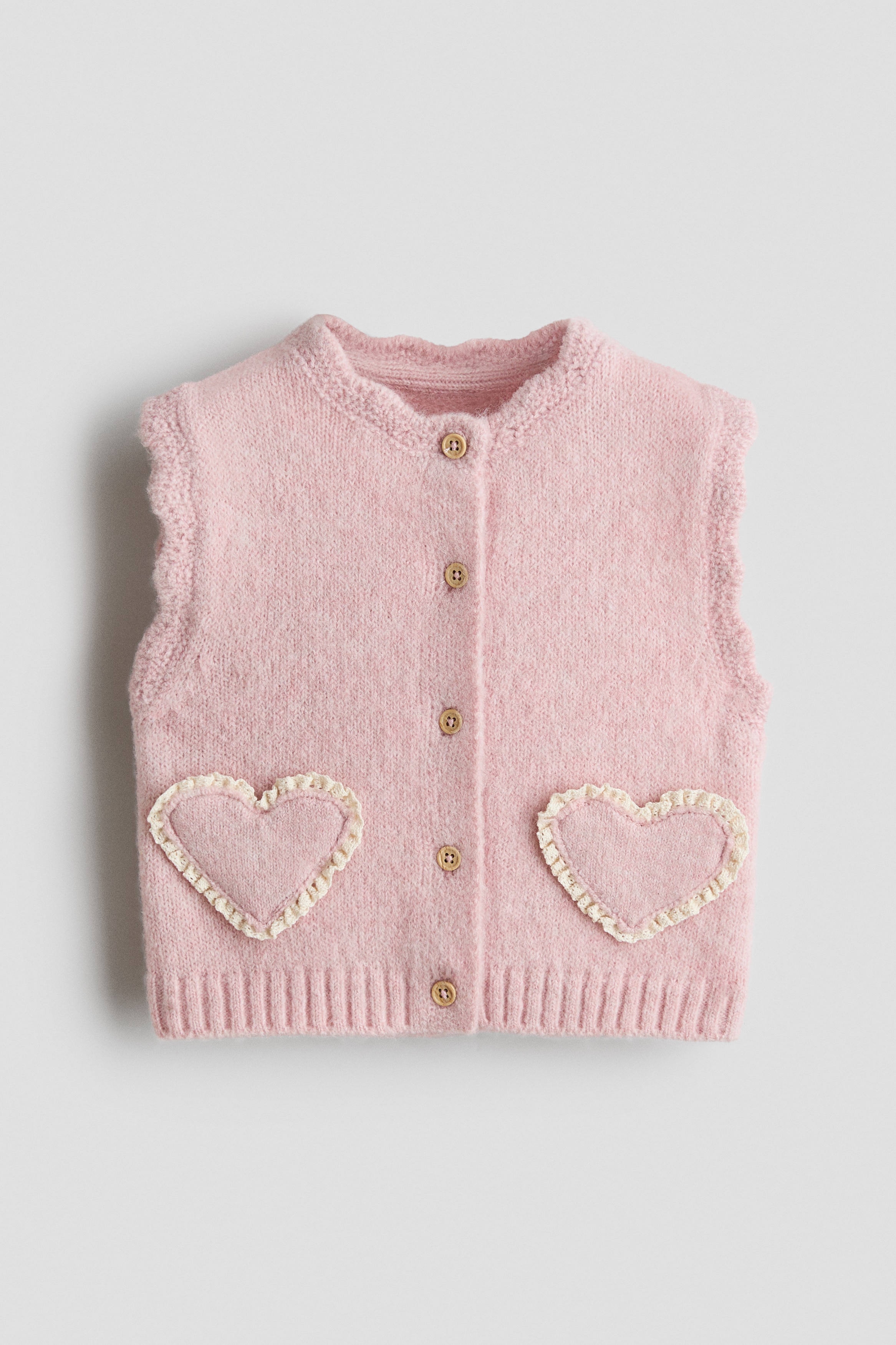 A light pink knitted vest is shown from the front, featuring a round scalloped neckline, a button-down front, and scalloped edges around the armholes. The vest features two heart-shaped front pockets detailed with white lace trim and is layered over a long-sleeved cream top.