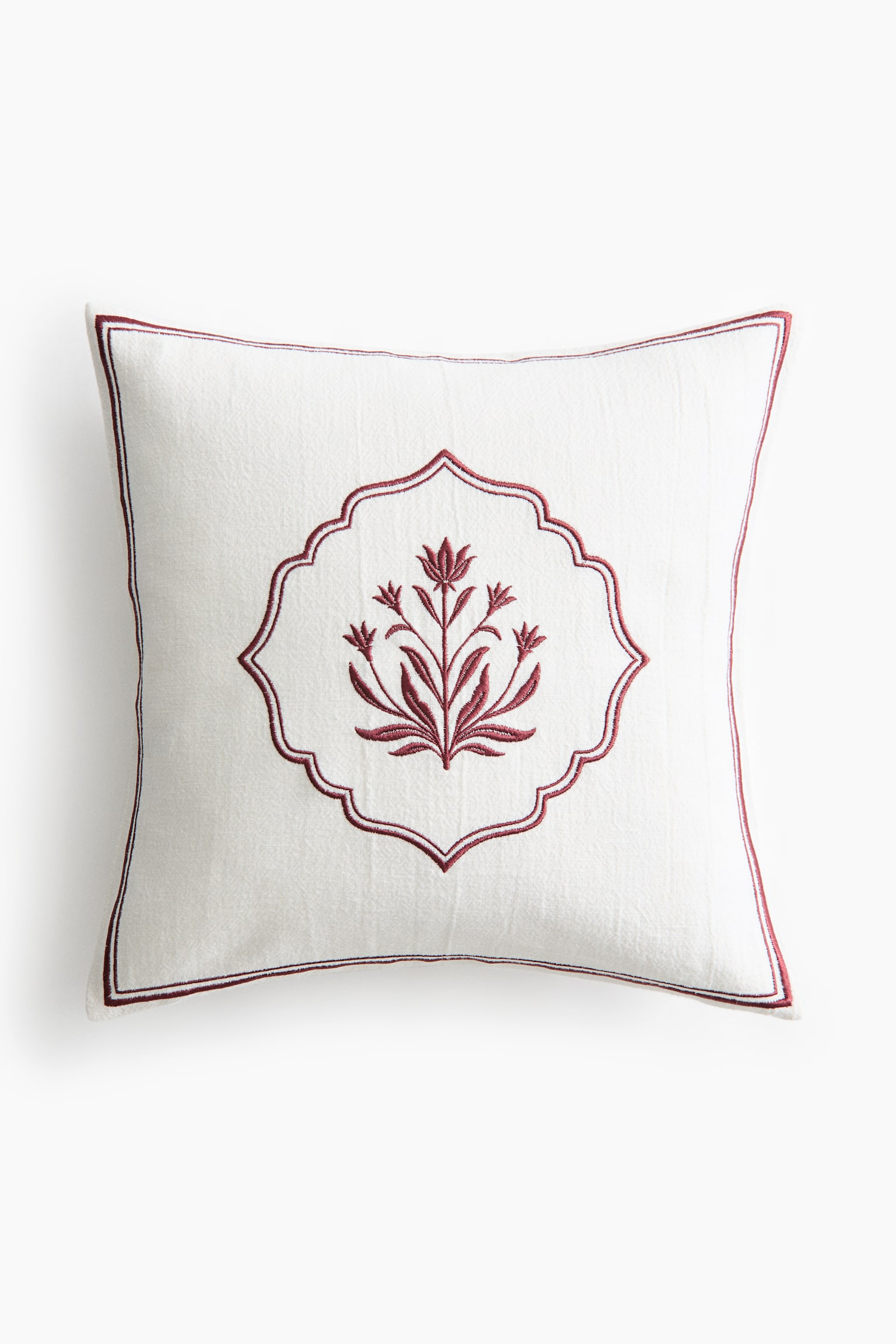 A square off-white decorative cushion is facing front, displaying a central dark red embroidered floral motif enclosed within a scalloped frame. A thin dark red embroidered border frames the outer edges of the textured fabric.