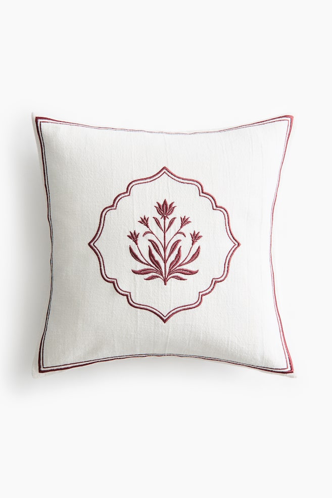 A square off-white decorative cushion is facing front, displaying a central dark red embroidered floral motif enclosed within a scalloped frame. A thin dark red embroidered border frames the outer edges of the textured fabric. - 1