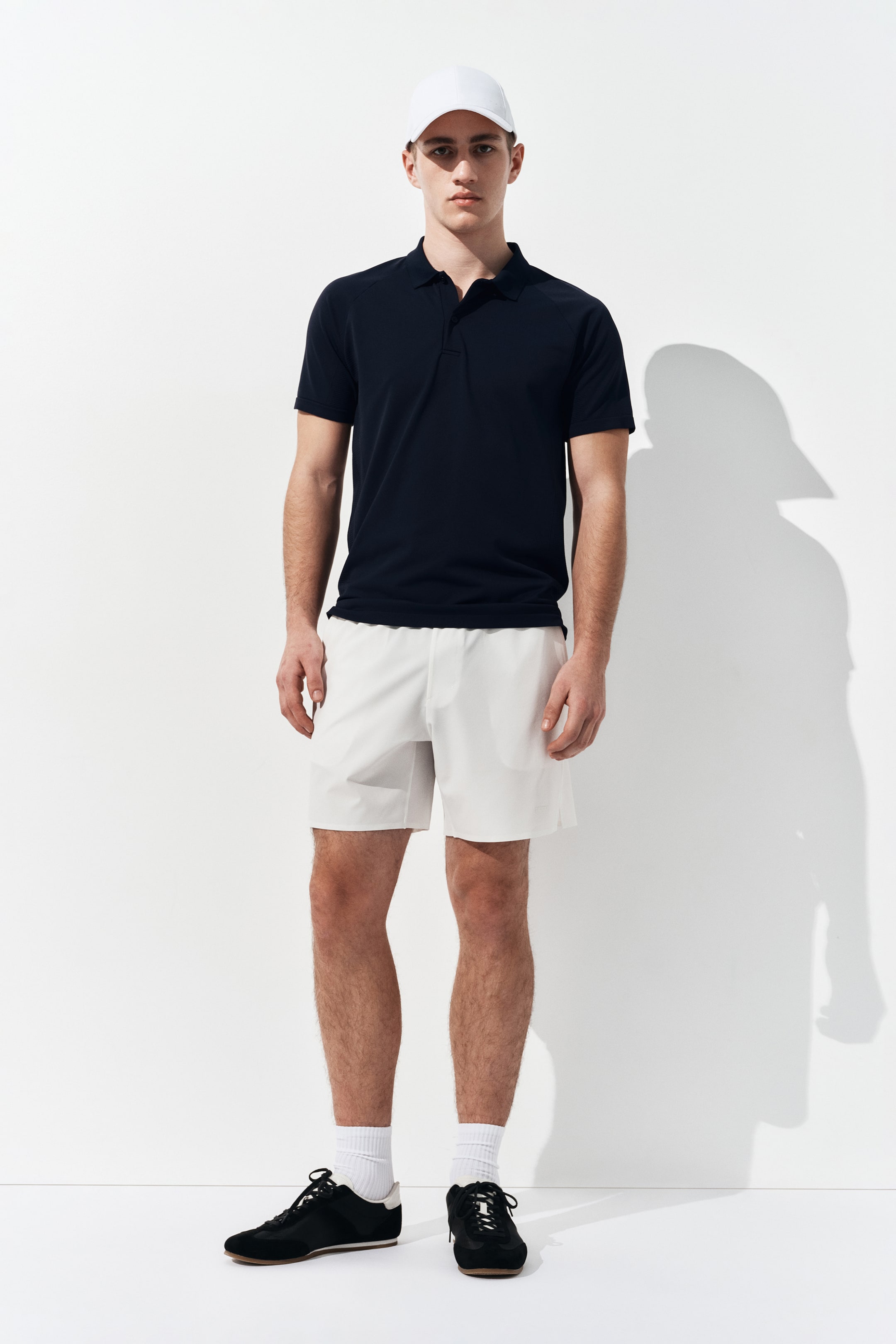 Agrandir l'image: La tenue complète est présentée de face, mettant en avant un polo à manches courtes bleu marine foncé avec un col classique et une patte de boutonnage à trois boutons. Elle est associée à un short de sport blanc cassé, une casquette de baseball blanche, des baskets basses noires et des chaussettes montantes blanches.