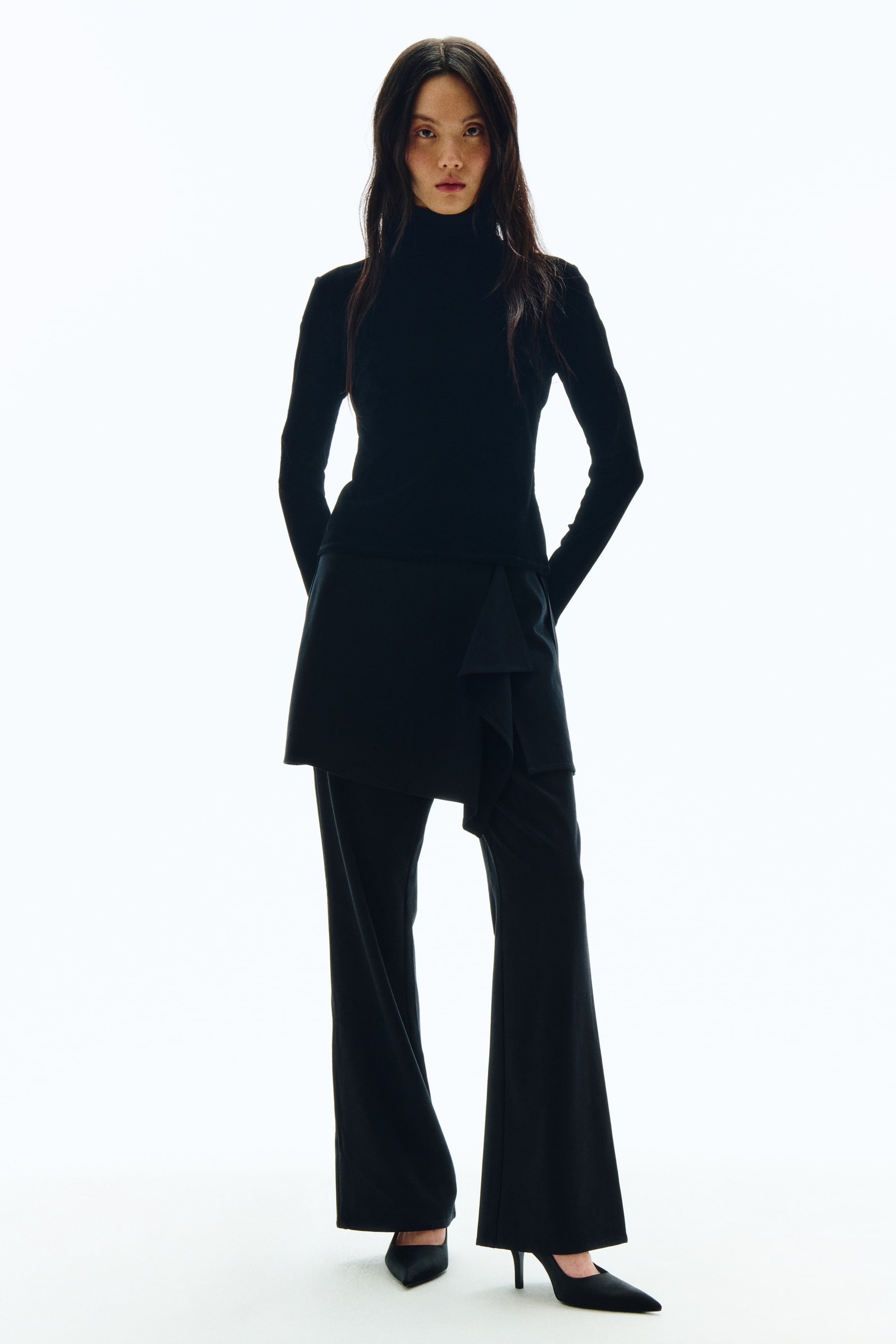 拡大画像を表示: ブラックの長袖タートルネックトップに、ブラックのフレアパンツを合わせたスタイリングをフロントアングルから捉えたカットです。右前身頃にドレープディテールをあしらったアシンメトリーなブラックのラップパネルがパンツの上にレイヤードされており、足元はブラックのポインテッドトゥヒールでコーディネートが完成しています。