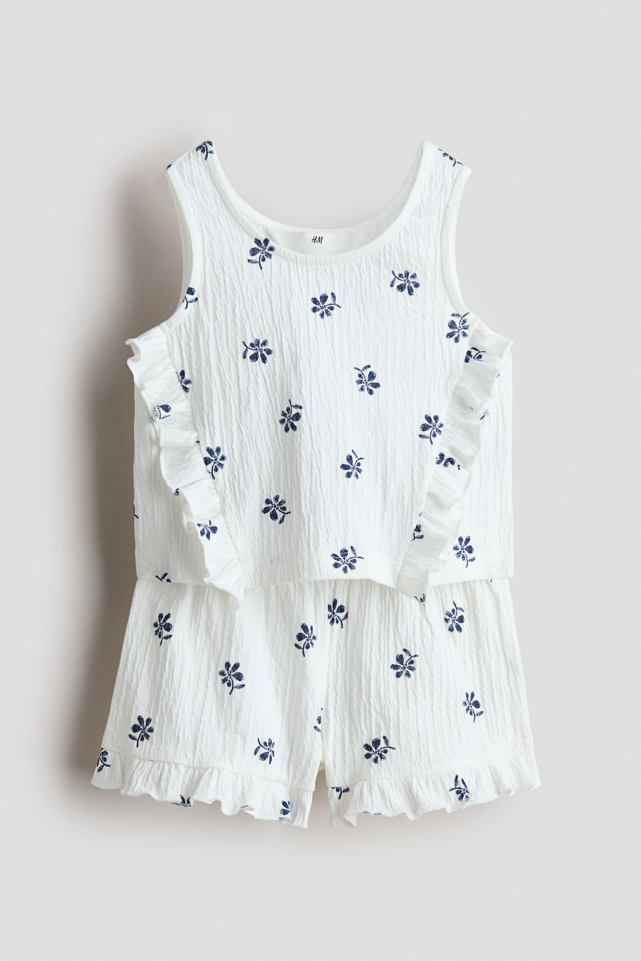 Grotere afbeelding bekijken: Een witte gekreukelde jersey set, van voren gezien, bestaat uit een hemdje en een short, beide versierd met een herhalend patroon van kleine blauwe bloemmotieven. Het hemdje is ontworpen met een ronde halslijn en ruches langs de voorkant, terwijl de short een elastische tailleband en ruches aan de zomen heeft.