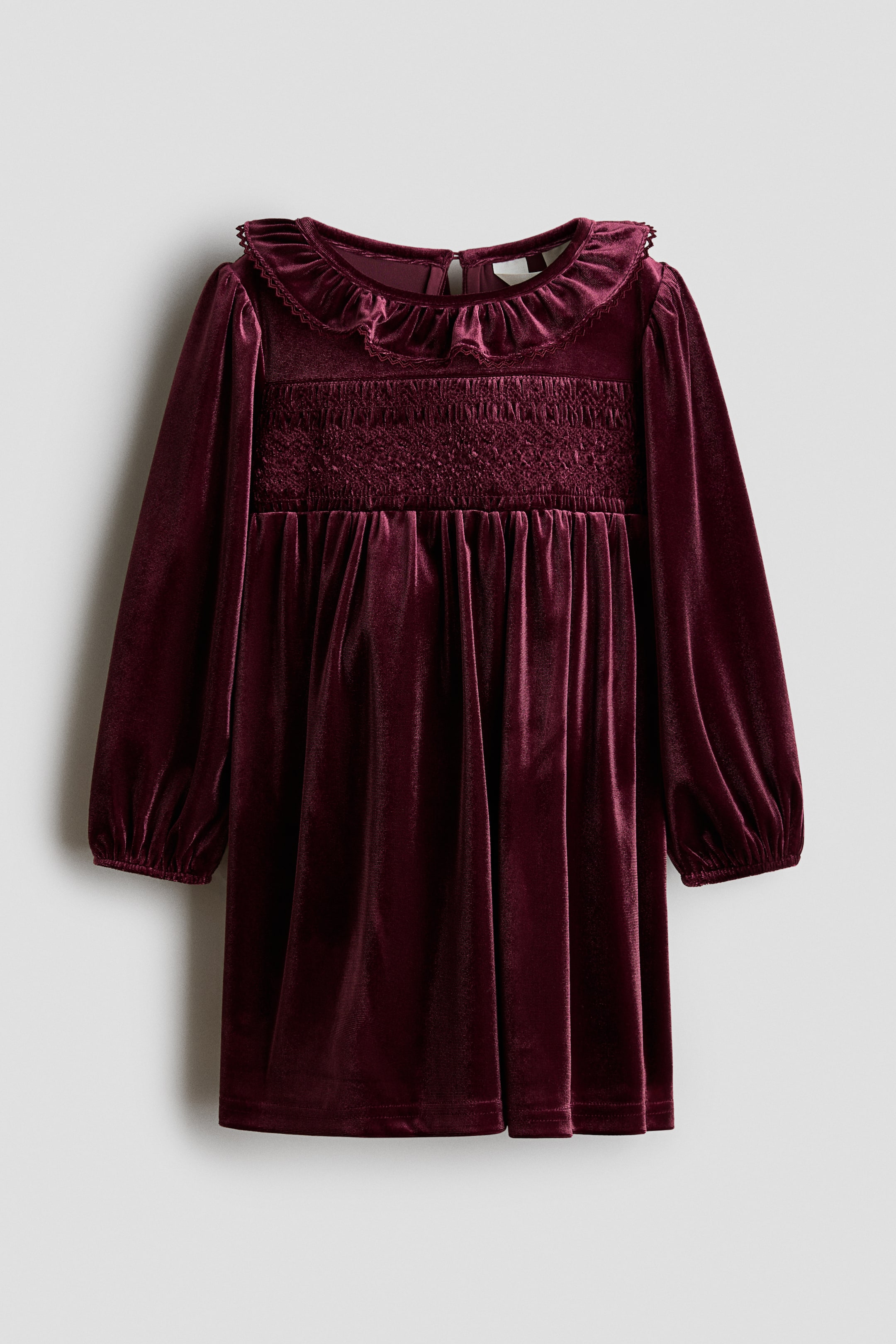 View larger image: A burgundy velvet dress is hanging front-facing, featuring a ruffled collar with lace trim and a smocked chest panel. It shows long sleeves with elasticated cuffs and a gathered skirt.