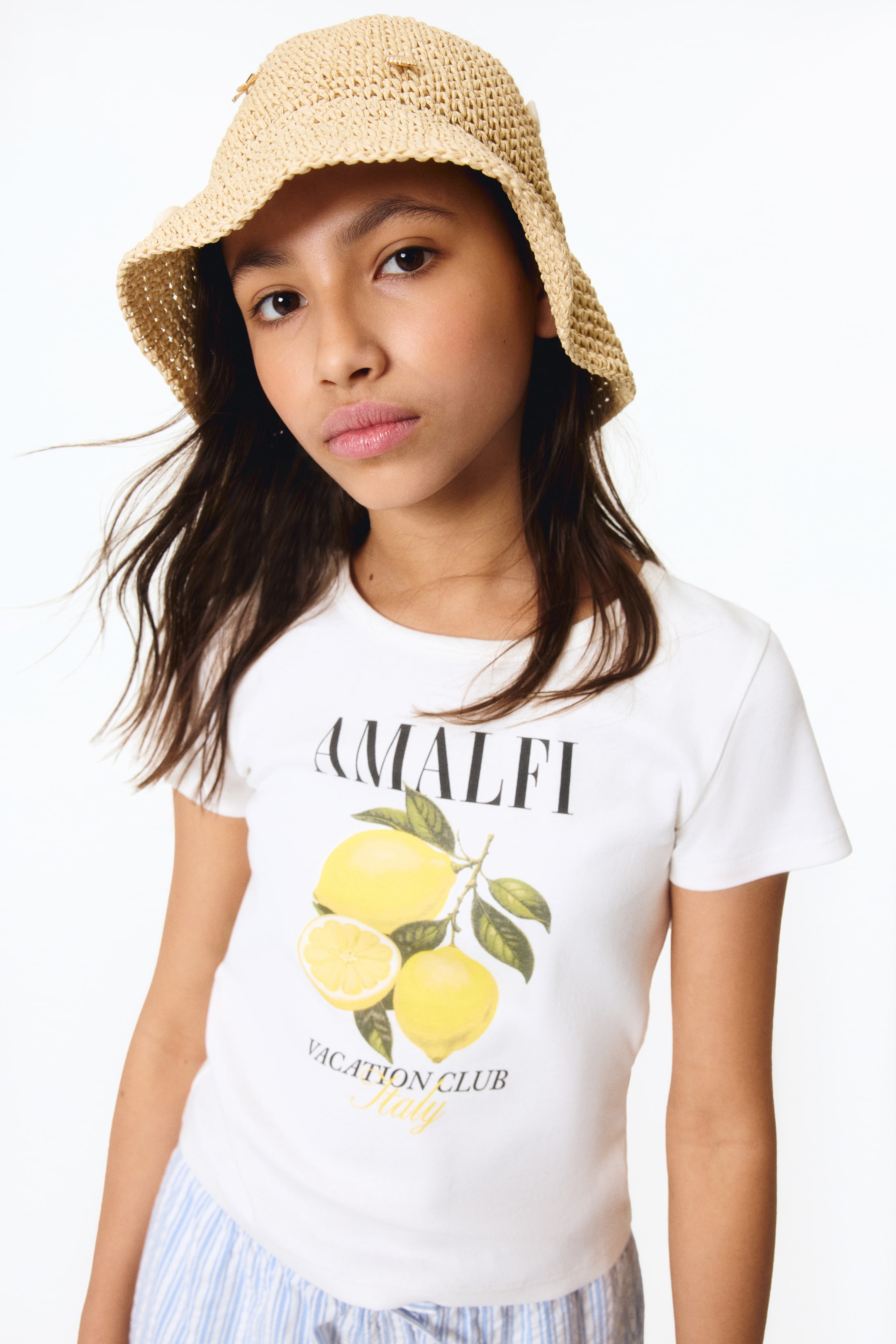 View larger image: A white T-shirt made from soft jersey features a ribbed trim around the neckline. The front showcases a graphic print with "AMALFI" above a cluster of lemons, accompanied by "VACATION CLUB Italy."