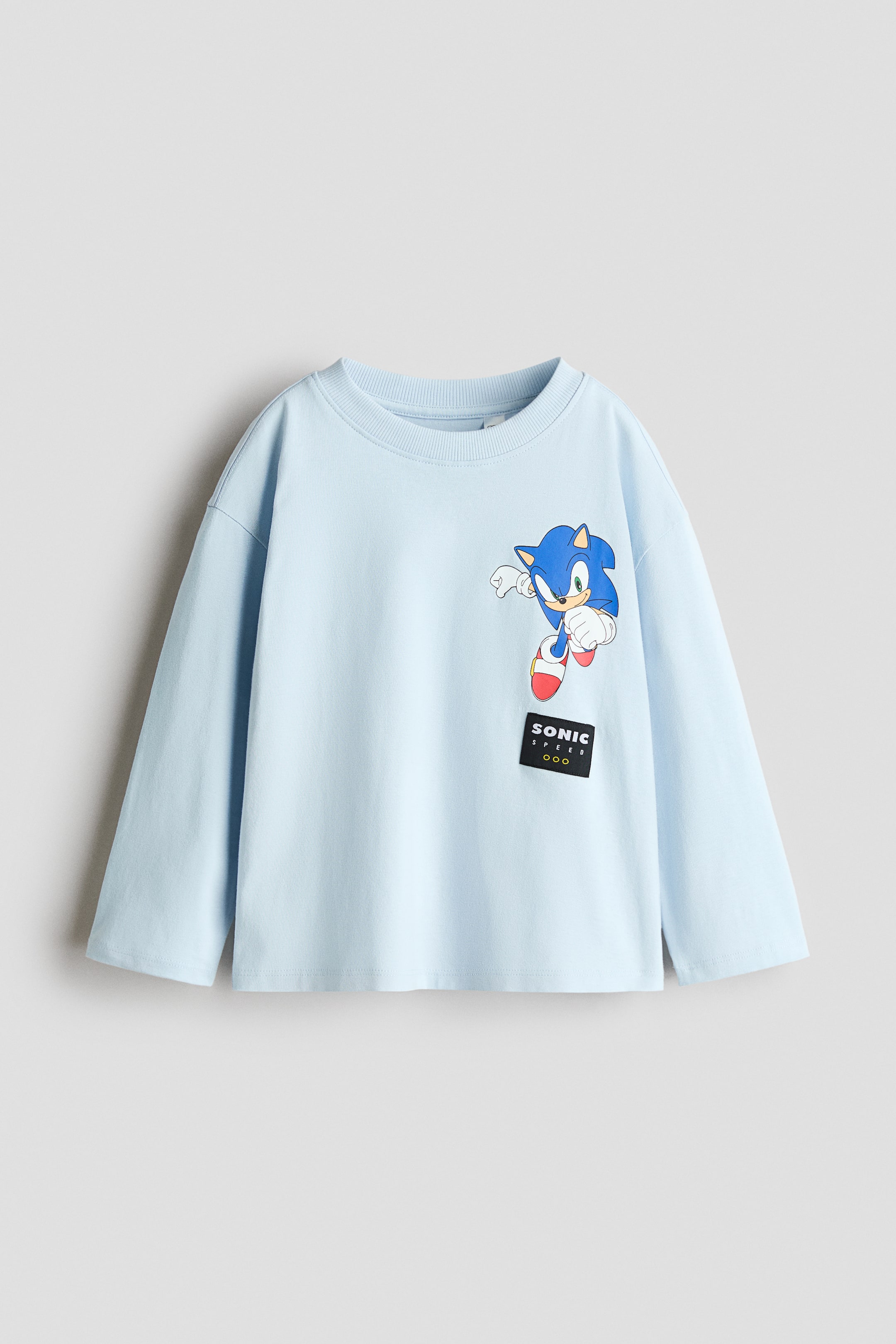 View larger image: A light blue loose-fit T-shirt is shown from the front, featuring a round rib-trimmed neckline, dropped shoulders, and long sleeves with a straight hem. The front displays a graphic print of Sonic the Hedgehog on the left chest, with a small black rectangular patch reading "SONIC SPEED" underneath.
