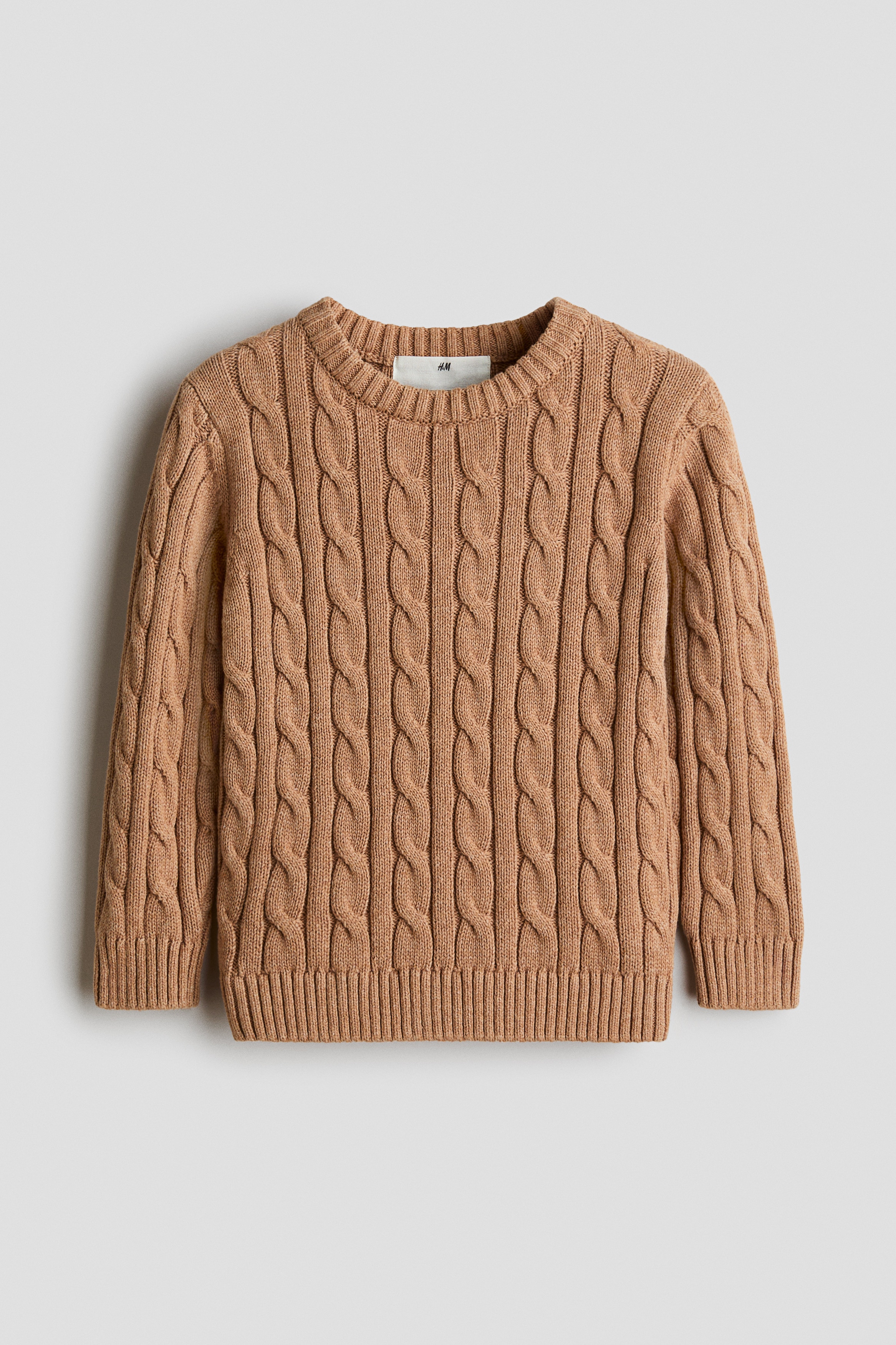 A light brown cable-knit cotton sweater is shown from the front, featuring a round ribbed neckline, ribbed cuffs, and a ribbed hem. The sweater displays a textured vertical cable-knit pattern across the body and sleeves.