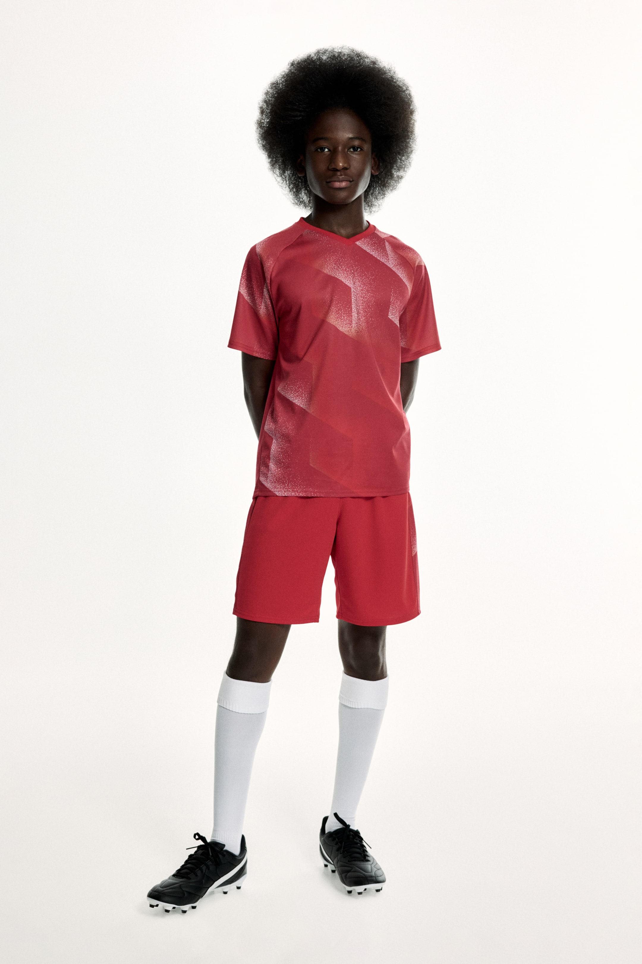 Agrandir l'image: Un haut de sport rouge et un short assorti sont présentés de face. Le haut à manches courtes est doté d’un col en V et présente un motif géométrique de couleur claire sur les épaules, les manches et le devant, coordonné au short uni rouge qui arrive au‑dessus du genou.