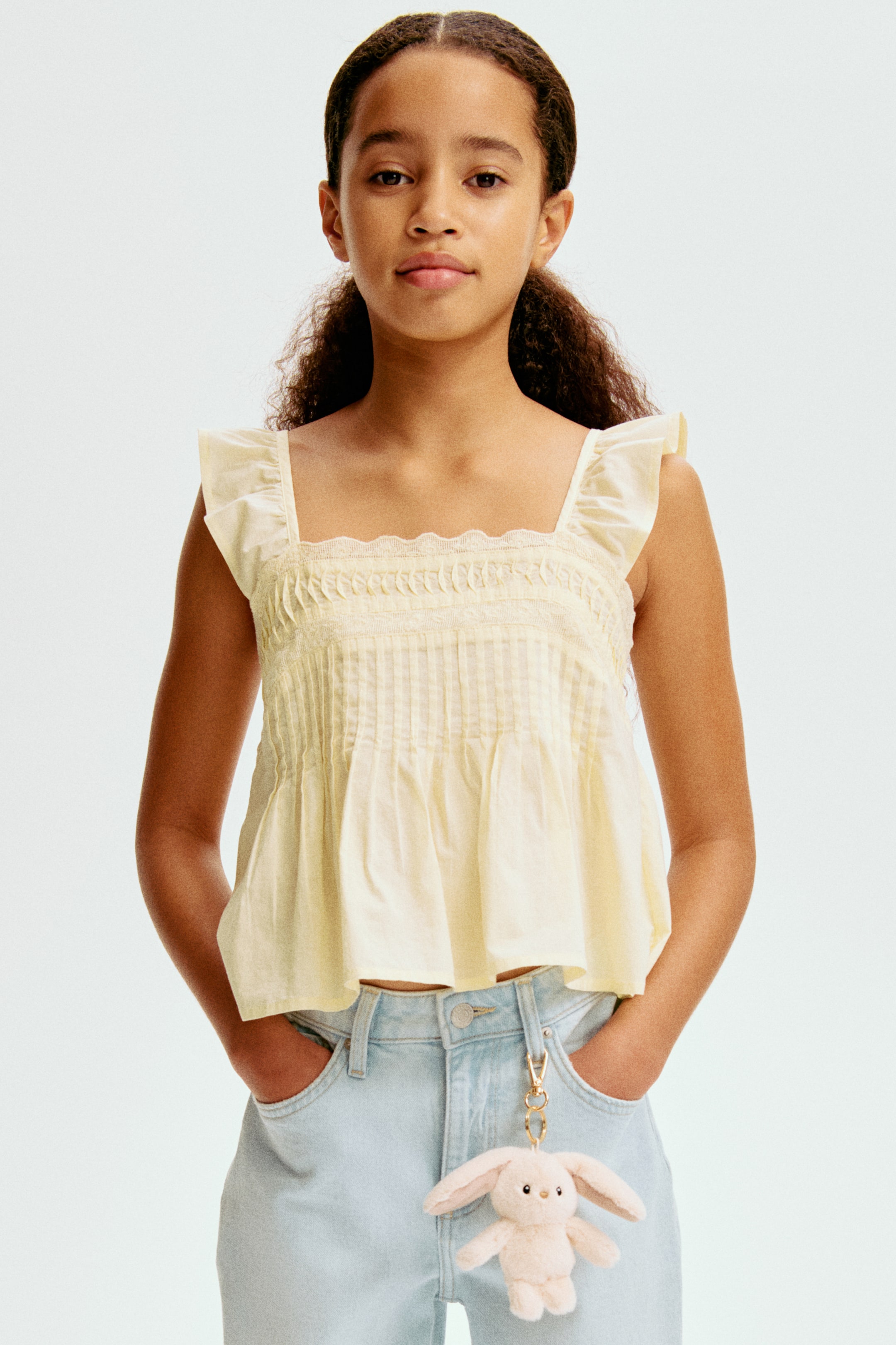 View larger image: The light yellow top features a square neckline adorned with a delicate trim and ruffled cap sleeves. The bodice is crafted with horizontal shirring across the bust and vertical pintucks, leading into a gathered, cropped hem.