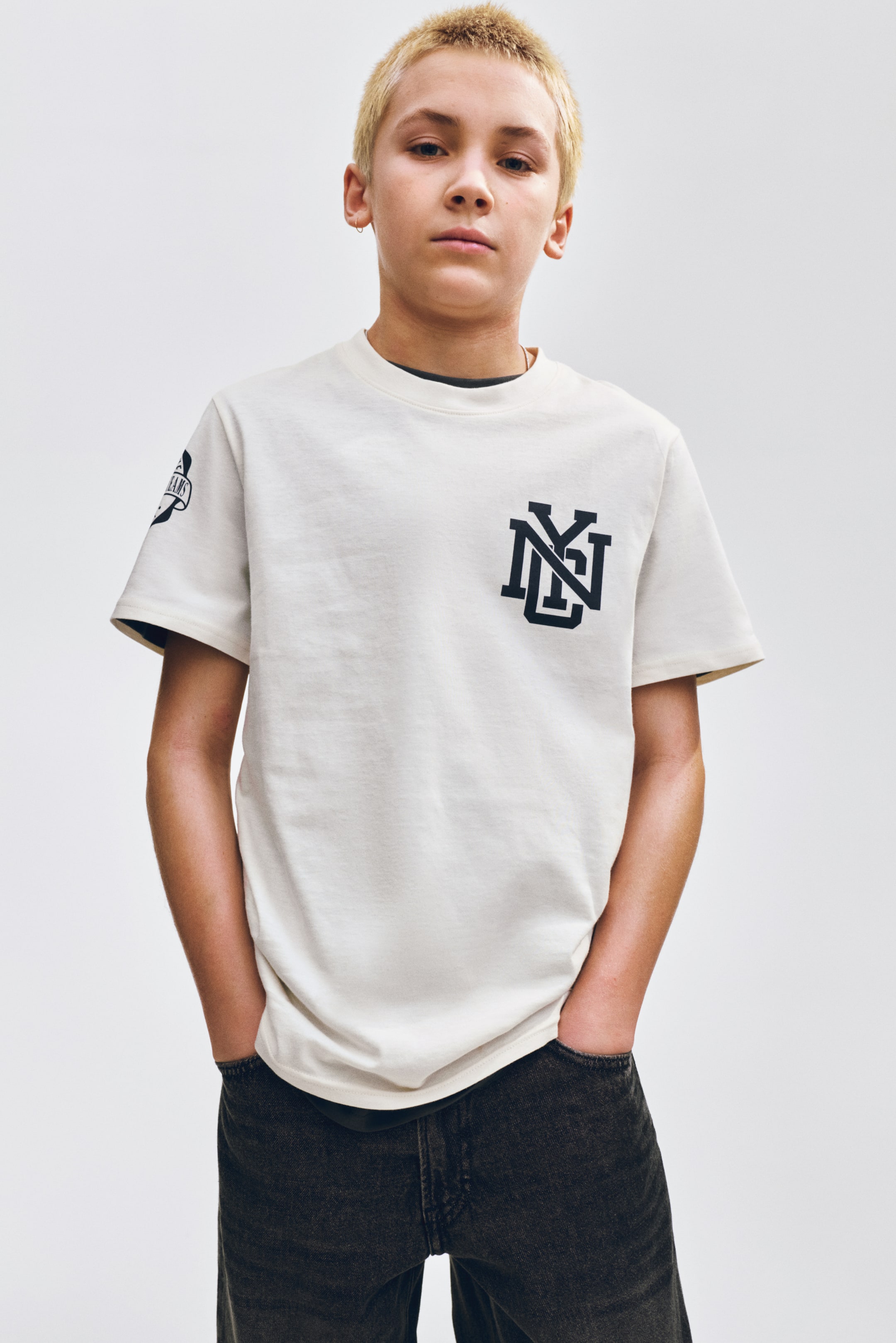 Visa större bild: En krämfärgad t-shirt med ett svart stiliserat "NY"-monogram på bröstet och en "DREAMS"-banderollgrafik på vänster ärm. Den har en rund halsringning och korta ärmar, med en mörk undertröja synlig vid halsringningen och ärmsluten.