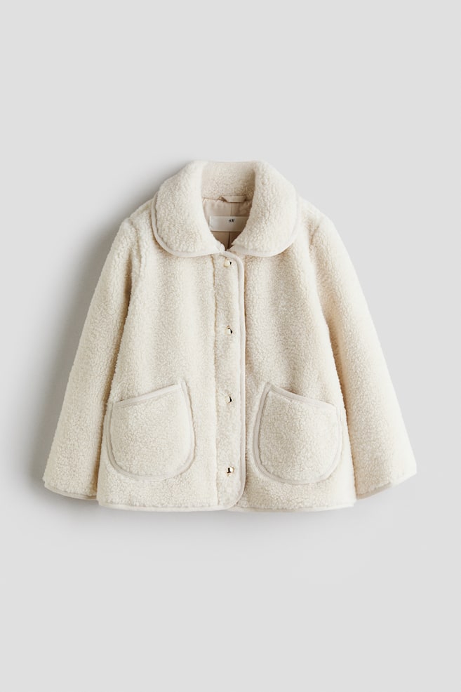 Een teddy fleece jas in crème kleur heeft een ronde kraag, een centrale knoopsluiting met goudkleurige knopen en twee grote, afgeronde opgestikte zakken aan de voorkant, allemaal gedetailleerd met een dun contrasterend biesje. - 1